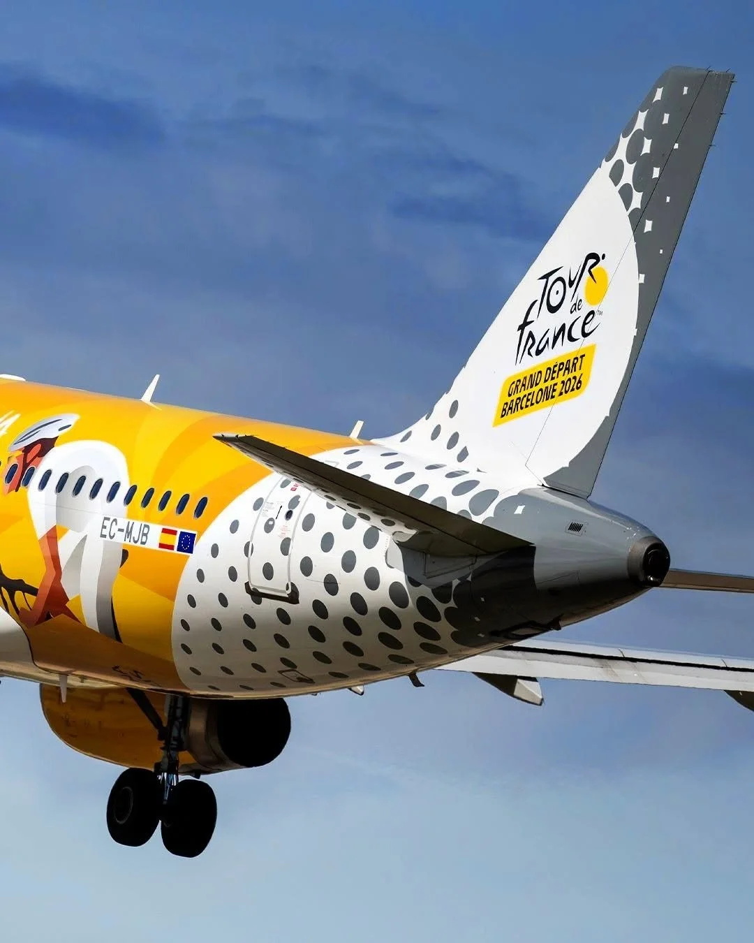 Close-up of a colorful Airplane with Tour de France livery, landing gear down, against a blue sky.