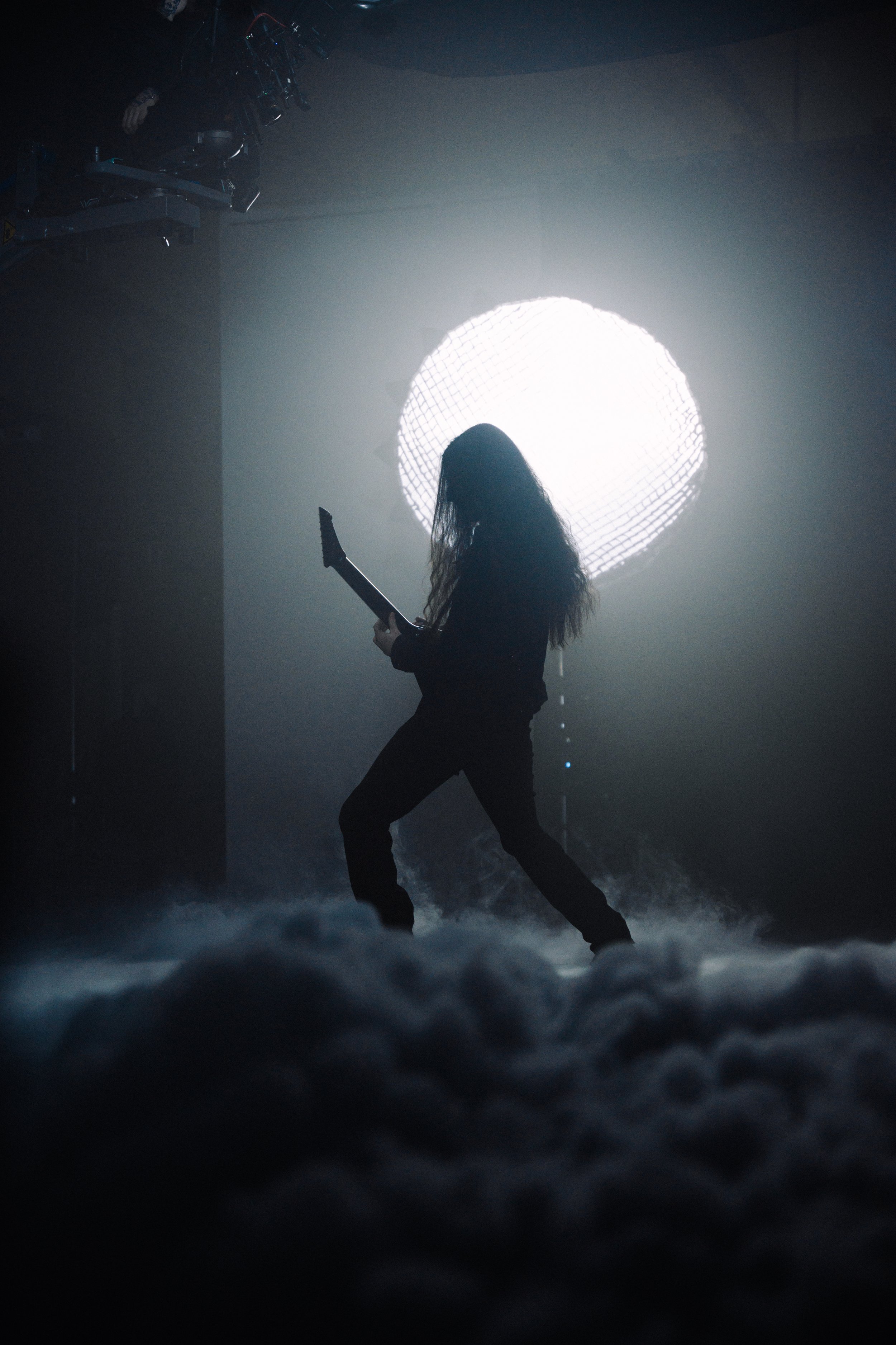Ein Musiker mit langer Frisur spielt Gitarre auf einer Bühne mit Rauch und hell erleuchteter Hintergrund, vermutlich während eines Live-Auftritts.