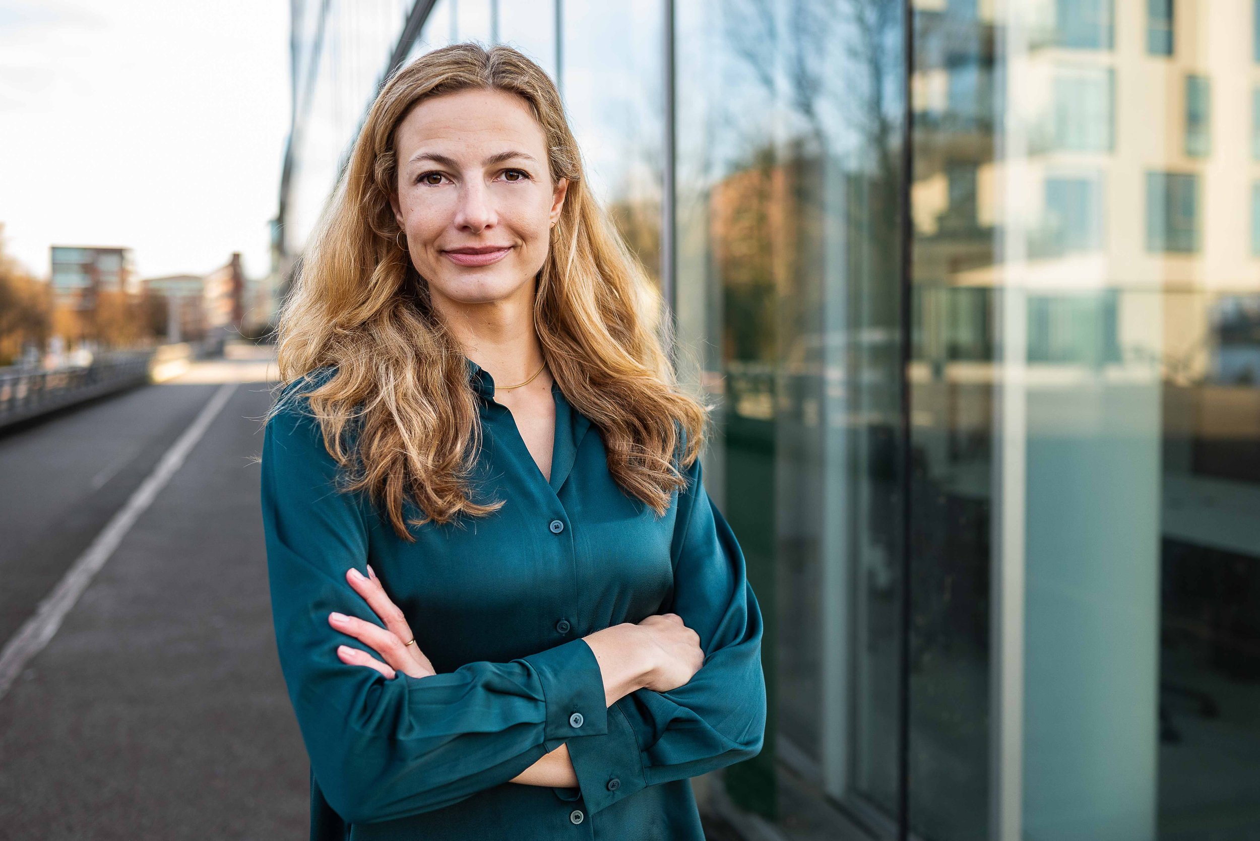 Sympathisches Business Portrait einer blonden Frau in blauer Bluse, am Hafen von Hamburg, freundlich lächelnd vor hellem Hintergrund.