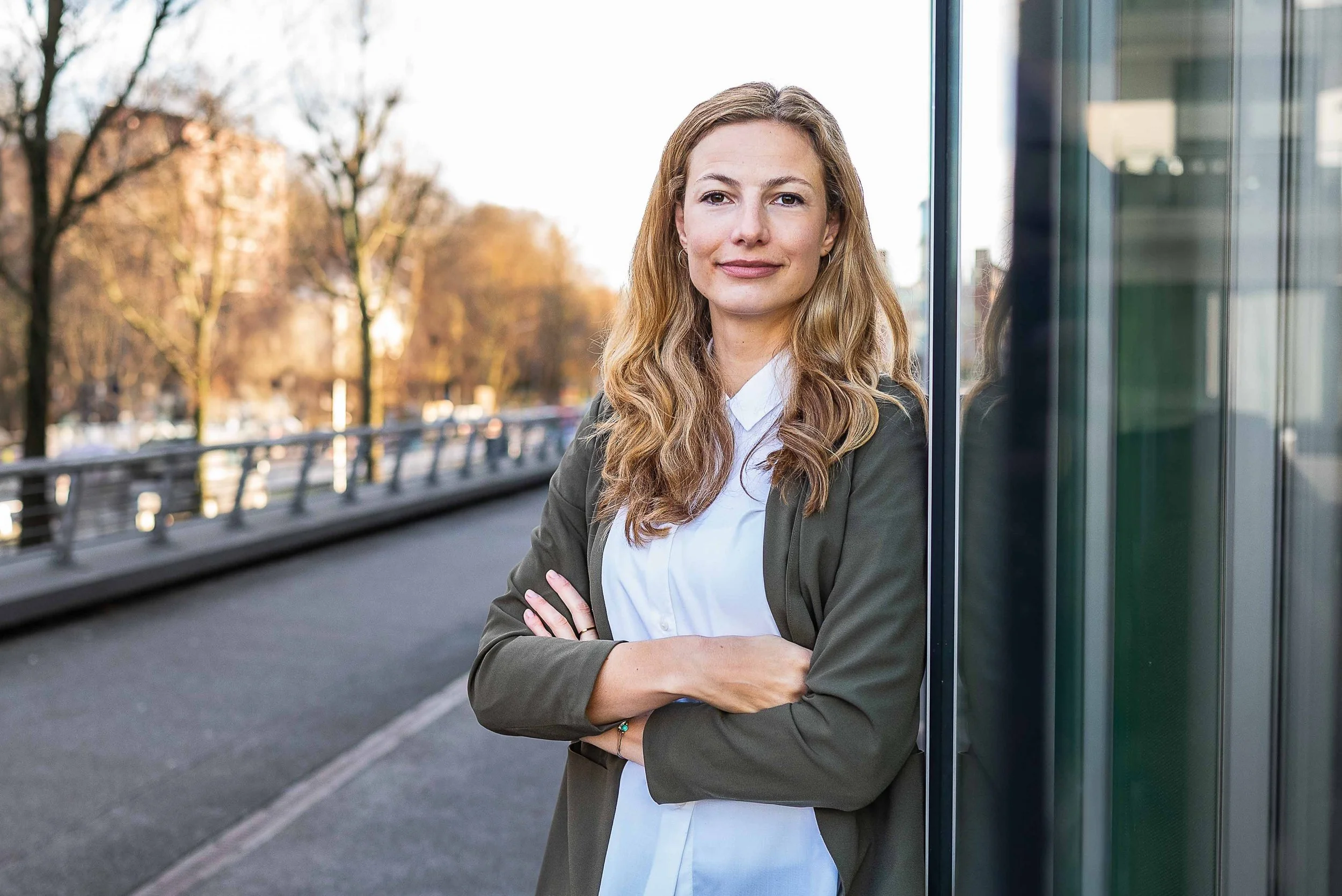Sympathisches Business Portrait einer blonden Frau im Blazer in Hamburg, natürlich lächelnd vor sonnigem Hintergrund.