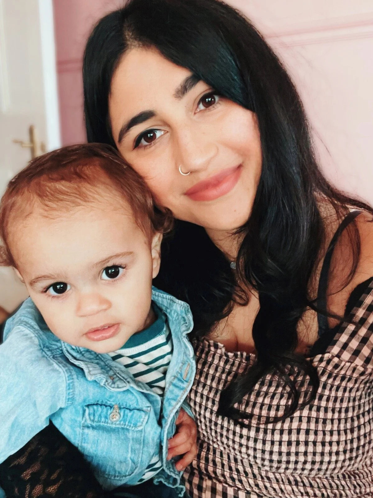 Close-up of a woman with dark hair and a septum piercing smiling and holding a young child with curly hair, tan skin, and large eyes, both looking into the camera.