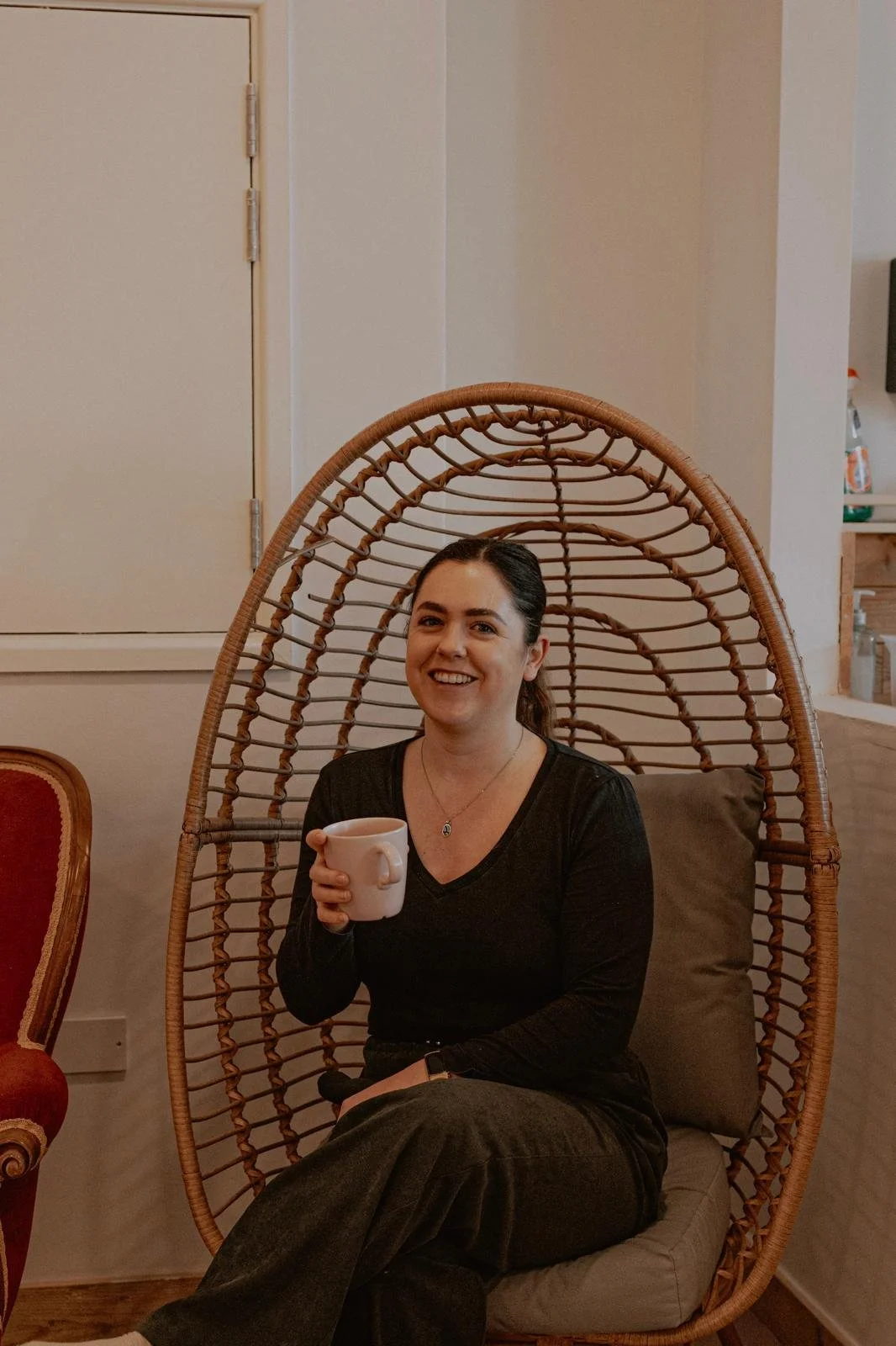 A woman smiling and holding a coffee mug while sitting in a wicker egg-shaped chair indoors.