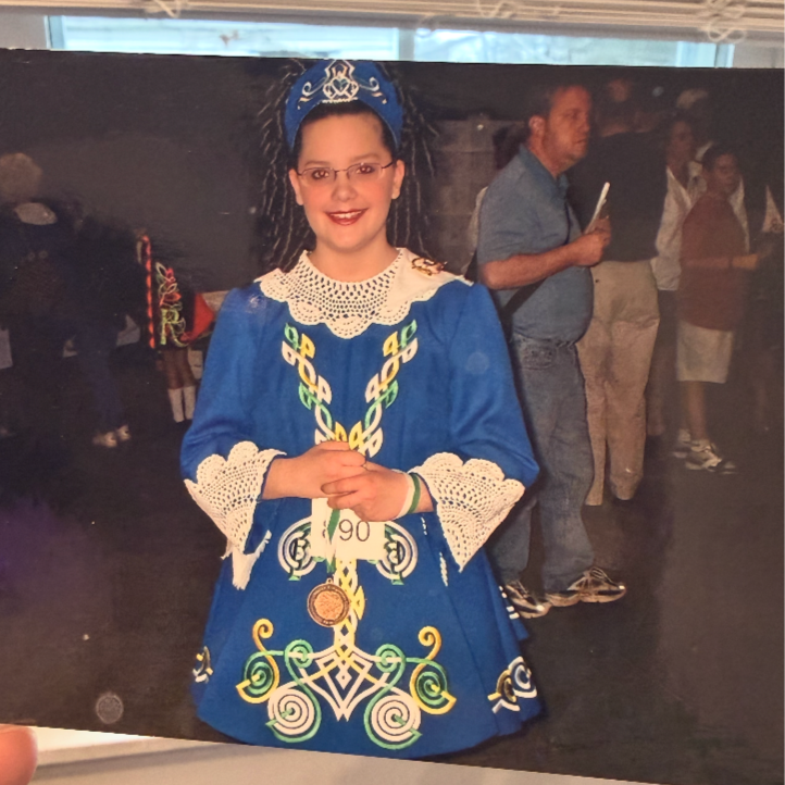 Young girl in a blue traditional dress with intricate embroidery and crochet details, wearing glasses and a matching headpiece, smiling and posing with her hands clasped.