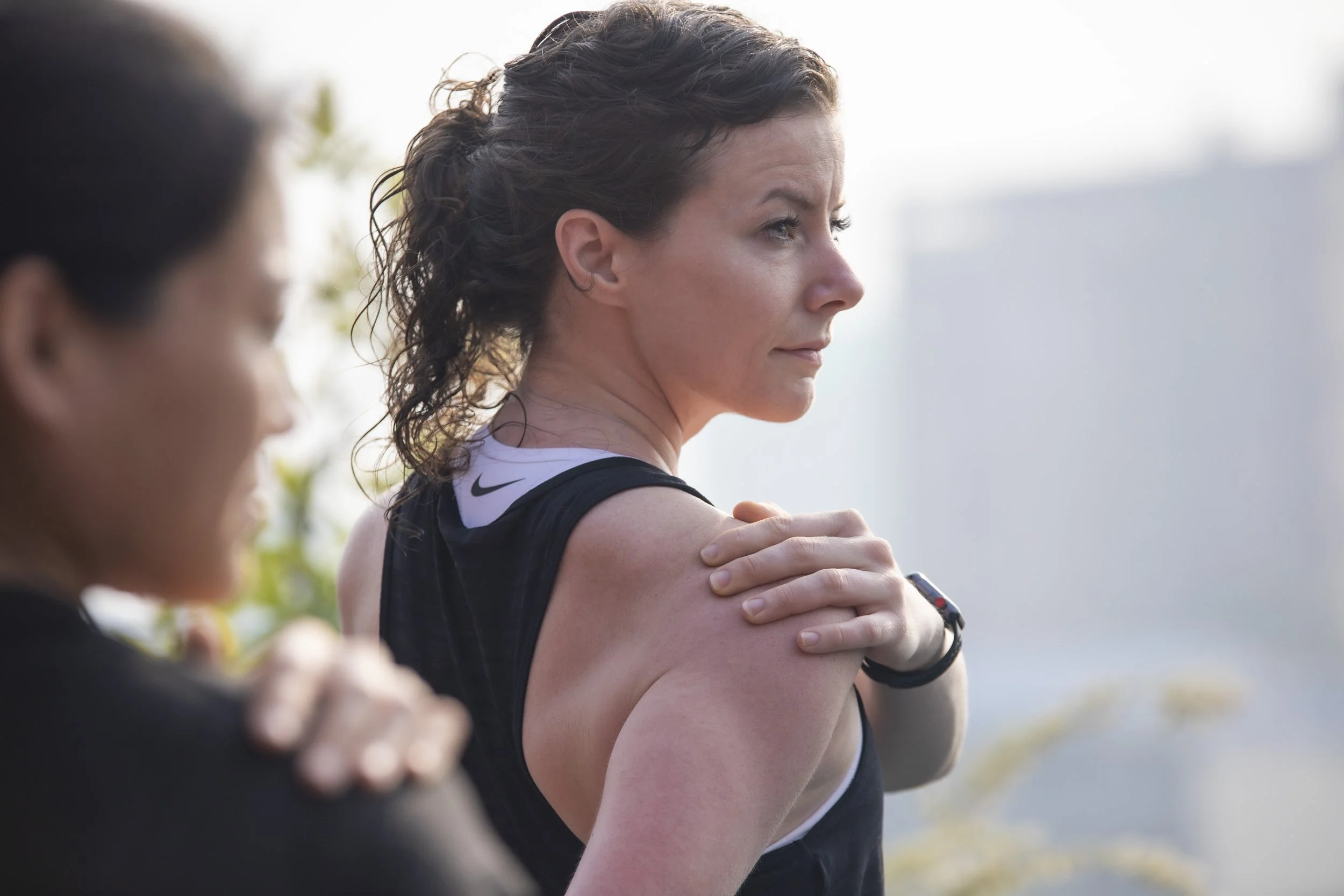 Two women at an outdoor fitness class, with one woman in focus stretching her shoulder while the other partially visible in the foreground.