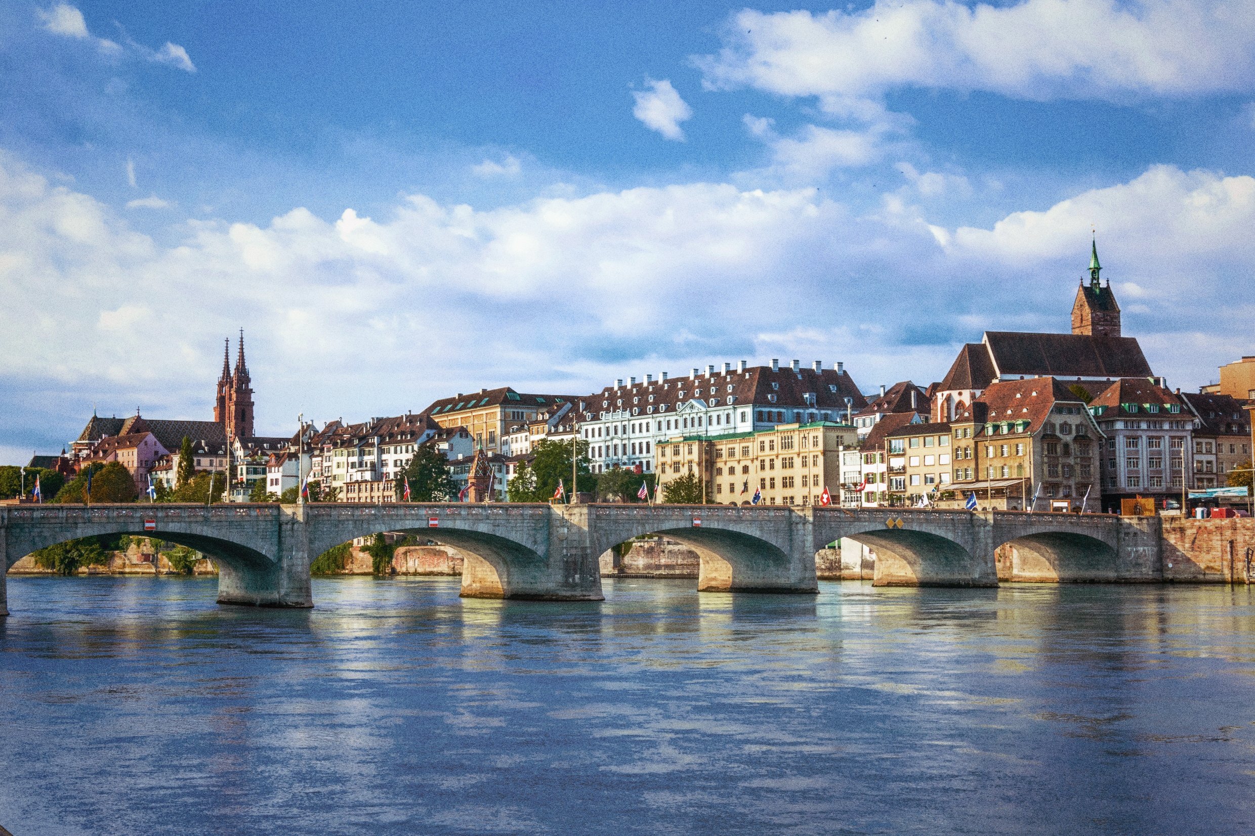 Cityscape with historic buildings, church steeples, and a stone bridge over a river under a partly cloudy sky.