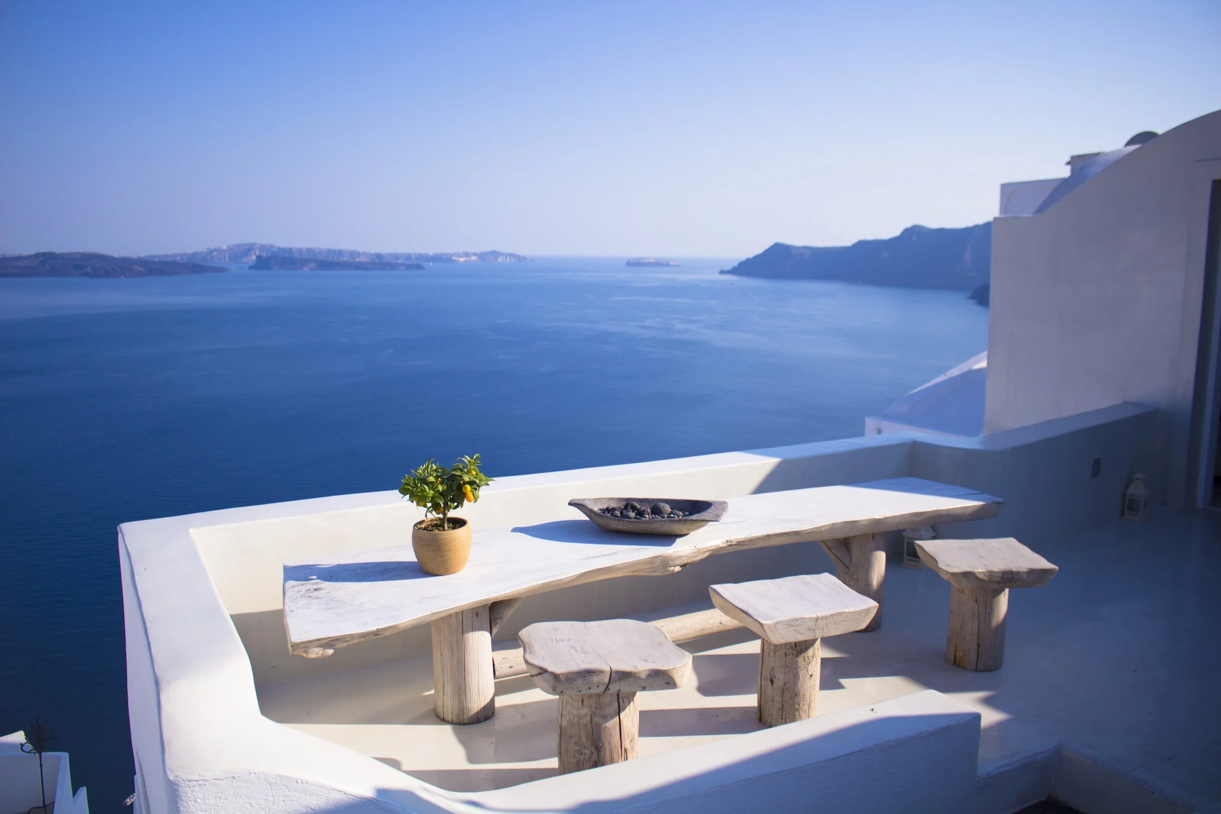 A white outdoor table with three matching stools, a small potted plant, and a dish with dark stones, overlooking a blue sea and distant islands in the background.