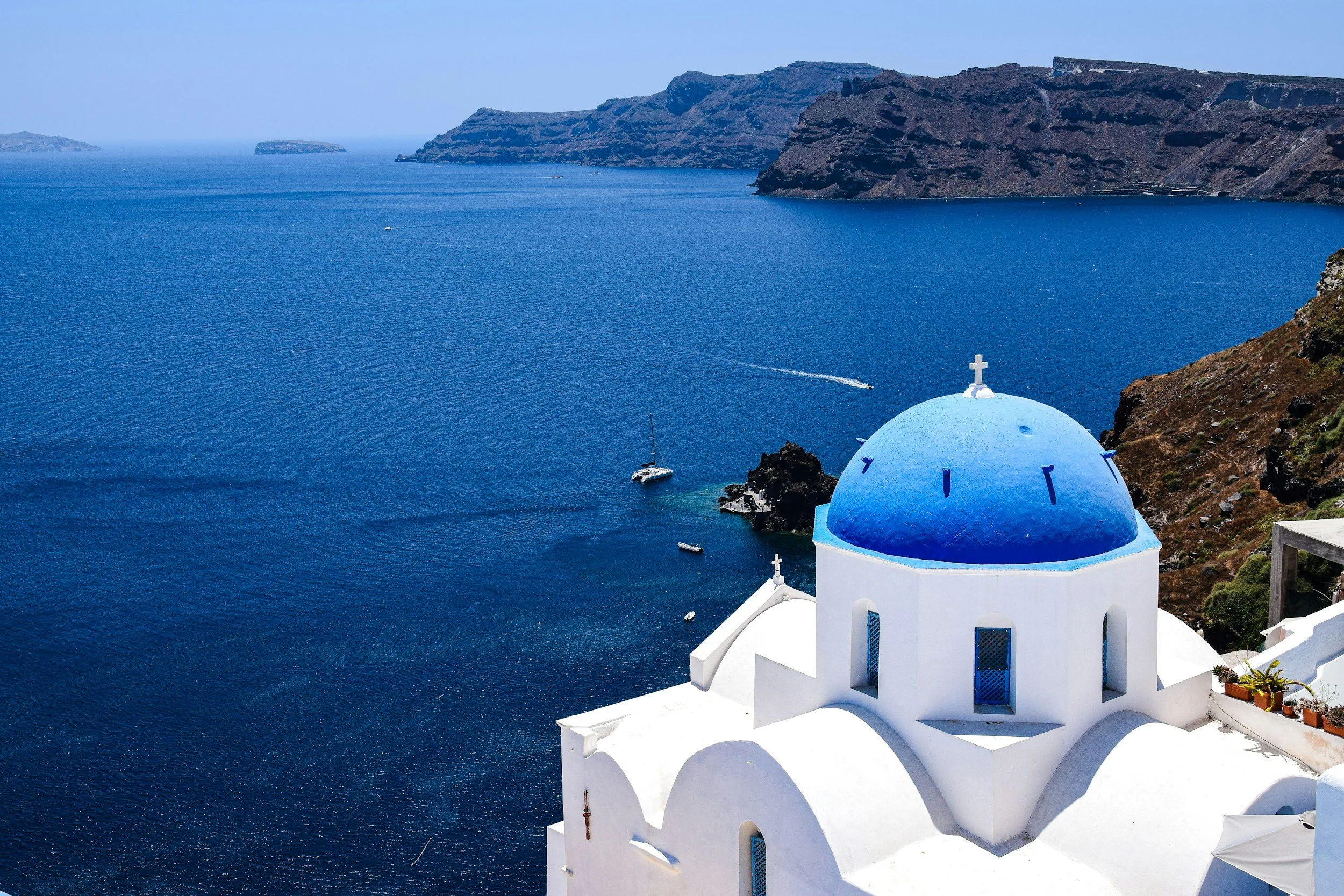 A white building with a blue dome roof and cross, overlooking the deep blue ocean with boats, rocky hills, and small islands in Santorini, Greece.