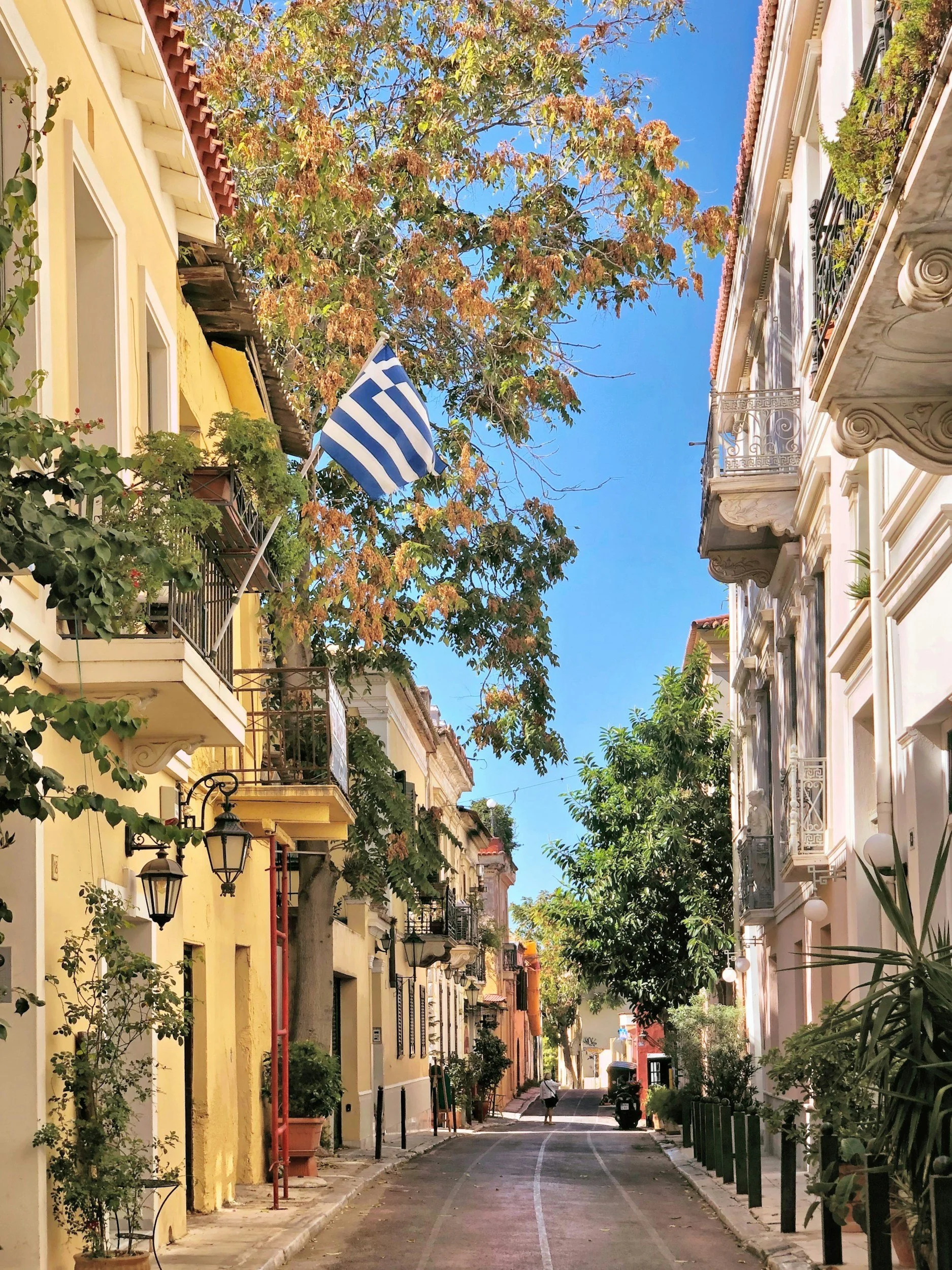 A narrow street in a European city with colorful buildings, balconies, and greenery. A Greek flag is flying from a balcony. The street is lined with plants, street lamps, and a person walking in the distance under a clear blue sky.