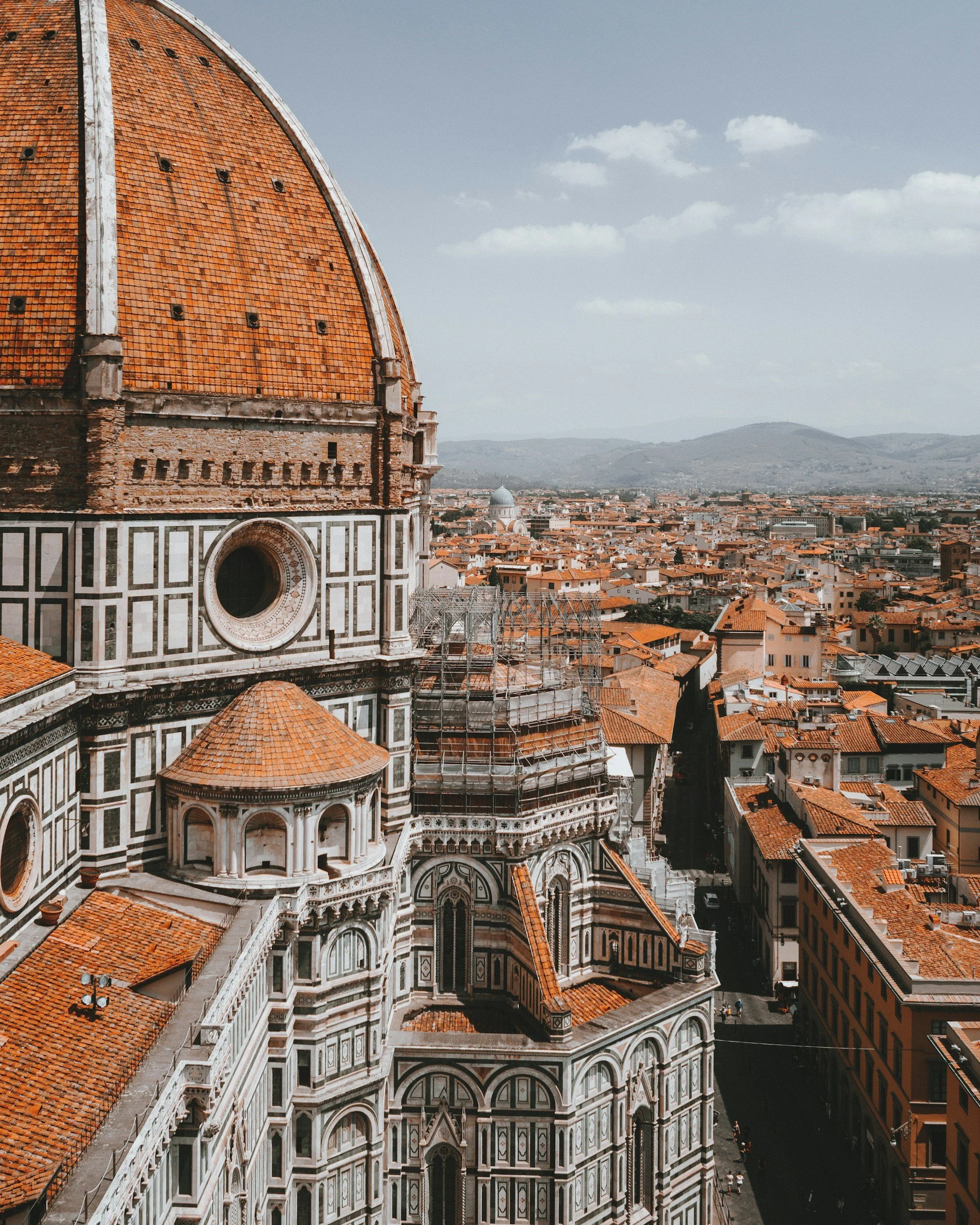 Aerial view of Florence, Italy showing the dome of Florence Cathedral (Duomo) with orange tiles, surrounded by historic buildings with orange roofs, and the rolling hills in the background.