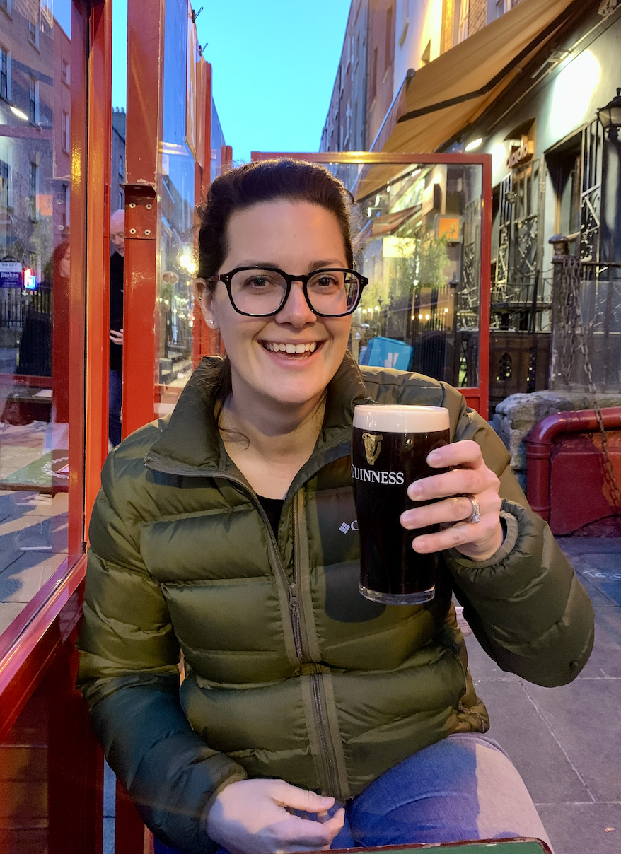 Hannah Clemente of Clemente Travels a Virtuoso Travel Advisor a woman smiling and holding a pint of Guinness beer at an outdoor pub or restaurant in the evening, wearing glasses and a green puffer jacket. Ireland travel specialist