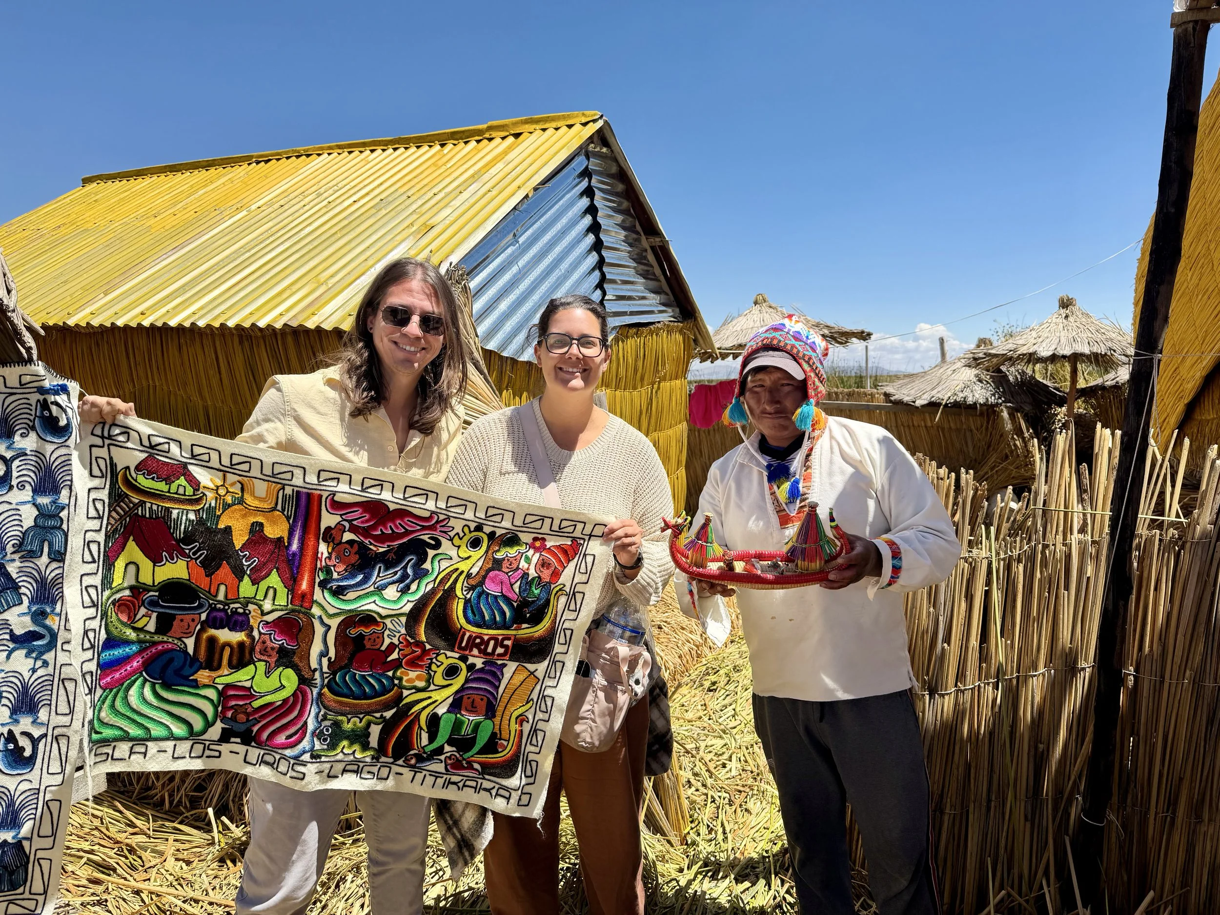 Three people standing outdoors with a thatched roof hut in the background, holding a colorful embroidered textile. The person on the left has long hair, sunglasses, and a white shirt. The person in the middle has glasses and a light sweater. The person on the right wears traditional indigenous attire including a headdress with tassels and is holding a small decorated item.