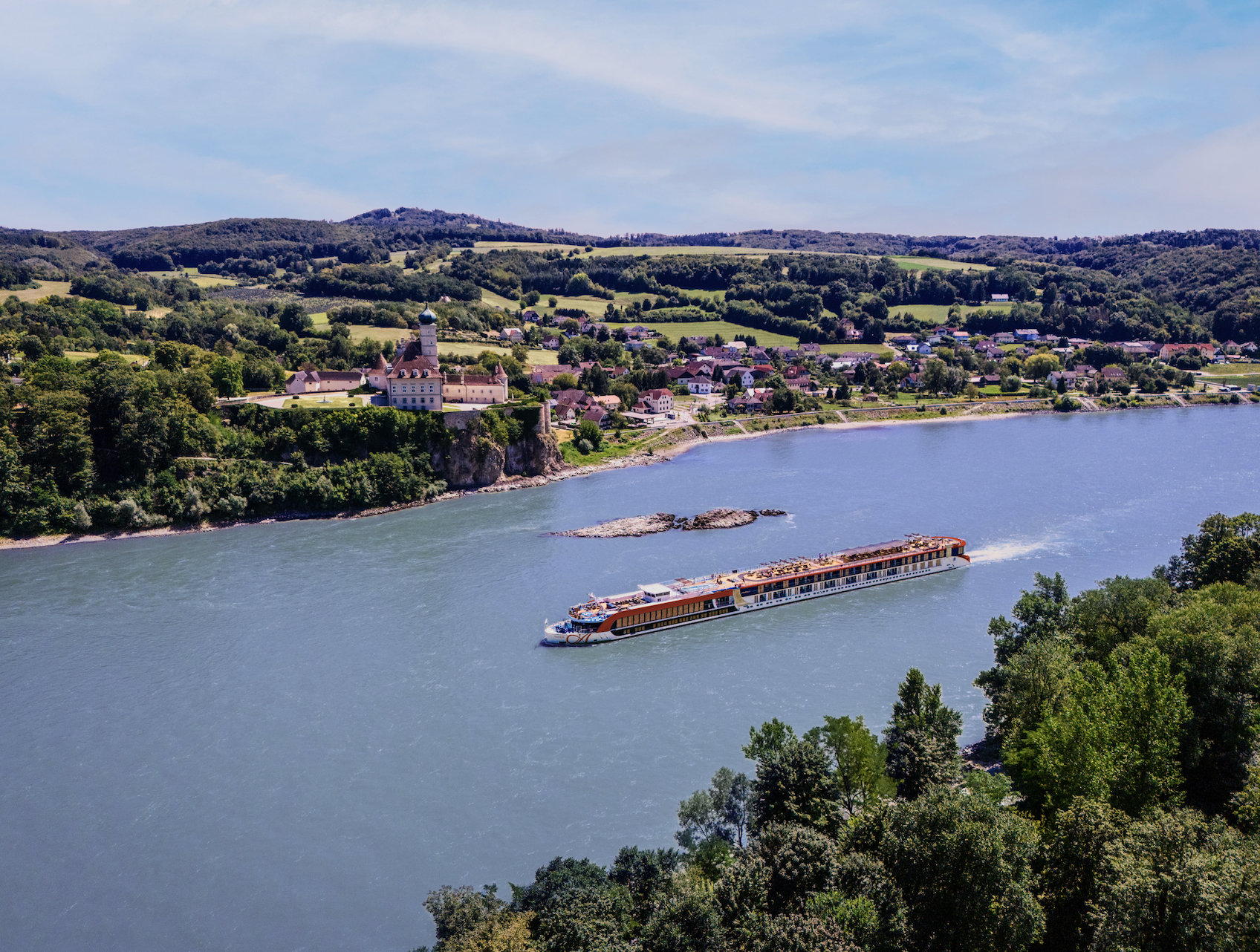 A scenic view of a river with a large boat sailing on it. Along the riverbank, there are green trees and a picturesque town with a castle and houses. The background features rolling hills under a partly cloudy sky.