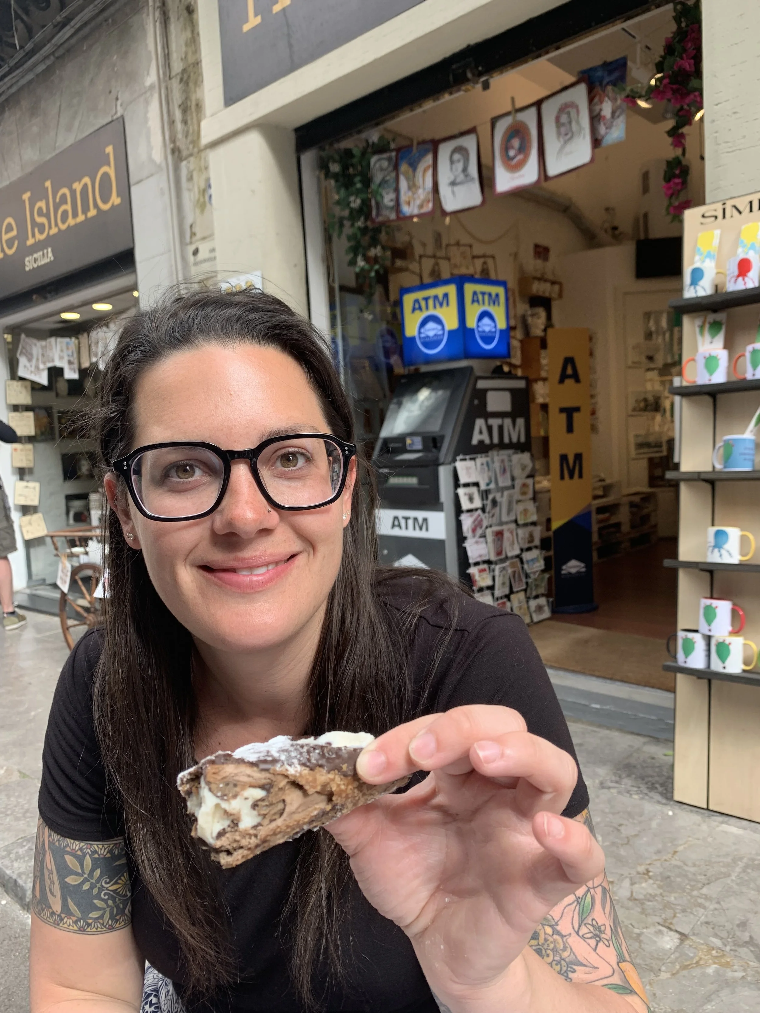 A woman with glasses and tattoos on her arms smiling and holding a large piece of ice cream or frosty dessert outdoors in front of a storefront with an ATM, greeting cards, and mugs on display.