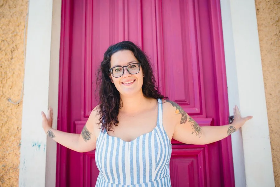 A smiling woman with dark curly hair, glasses, wearing a striped sleeveless dress, standing in front of a pink door, with arms outstretched, showing tattoos on her arms.