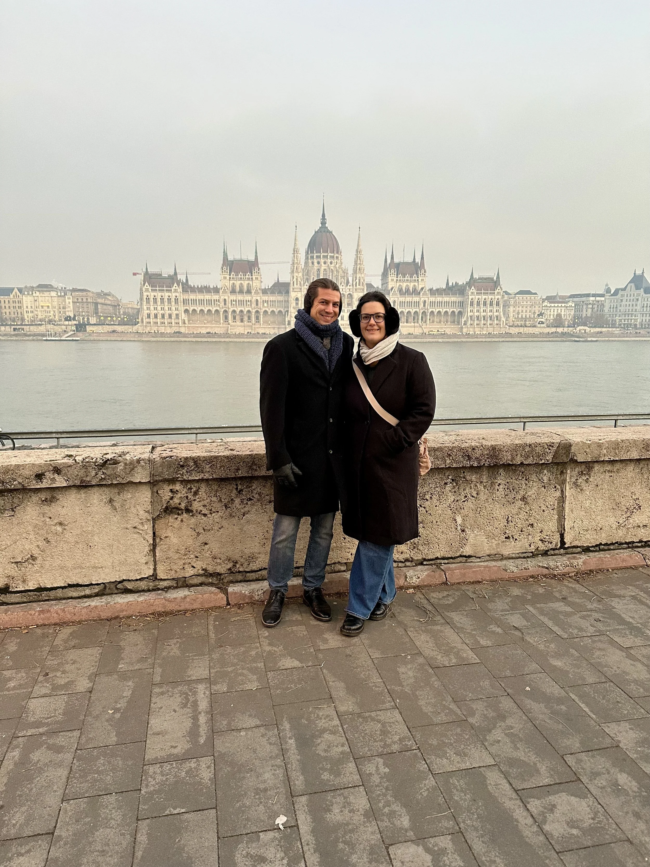 Hannah Clemente and Daniel Clemente of Clemente Travels a Virtuoso Travel Advisor, stand on a waterfront promenade with the Hungarian Parliament Building in Budapest in the background on a cloudy day. Massillon, Ohio travel advisor.