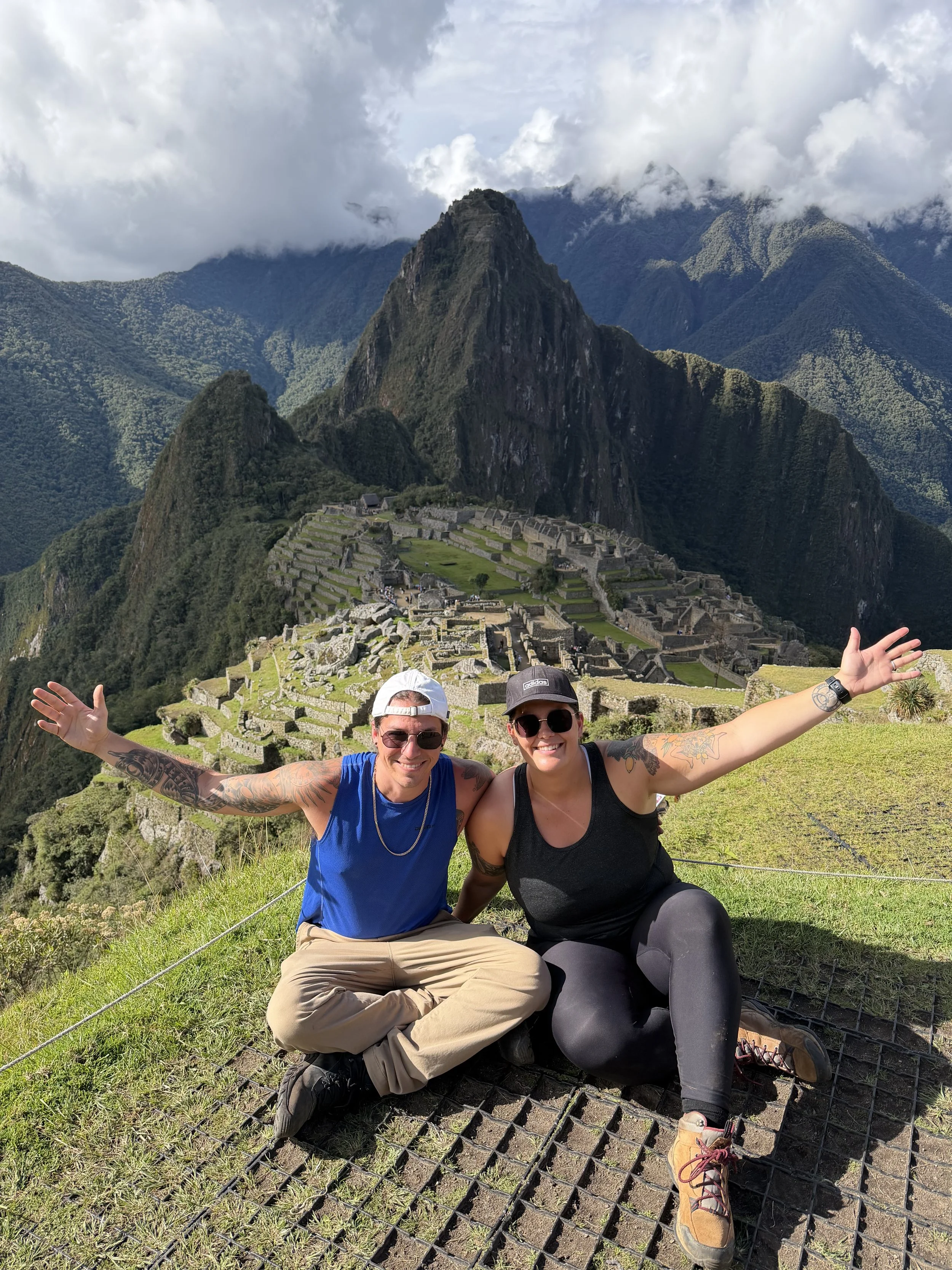 Two smiling women sitting on the ground with Machu Picchu and lush green mountains in the background. They are wearing sunglasses, baseball caps, and athletic clothing, with arms outstretched.