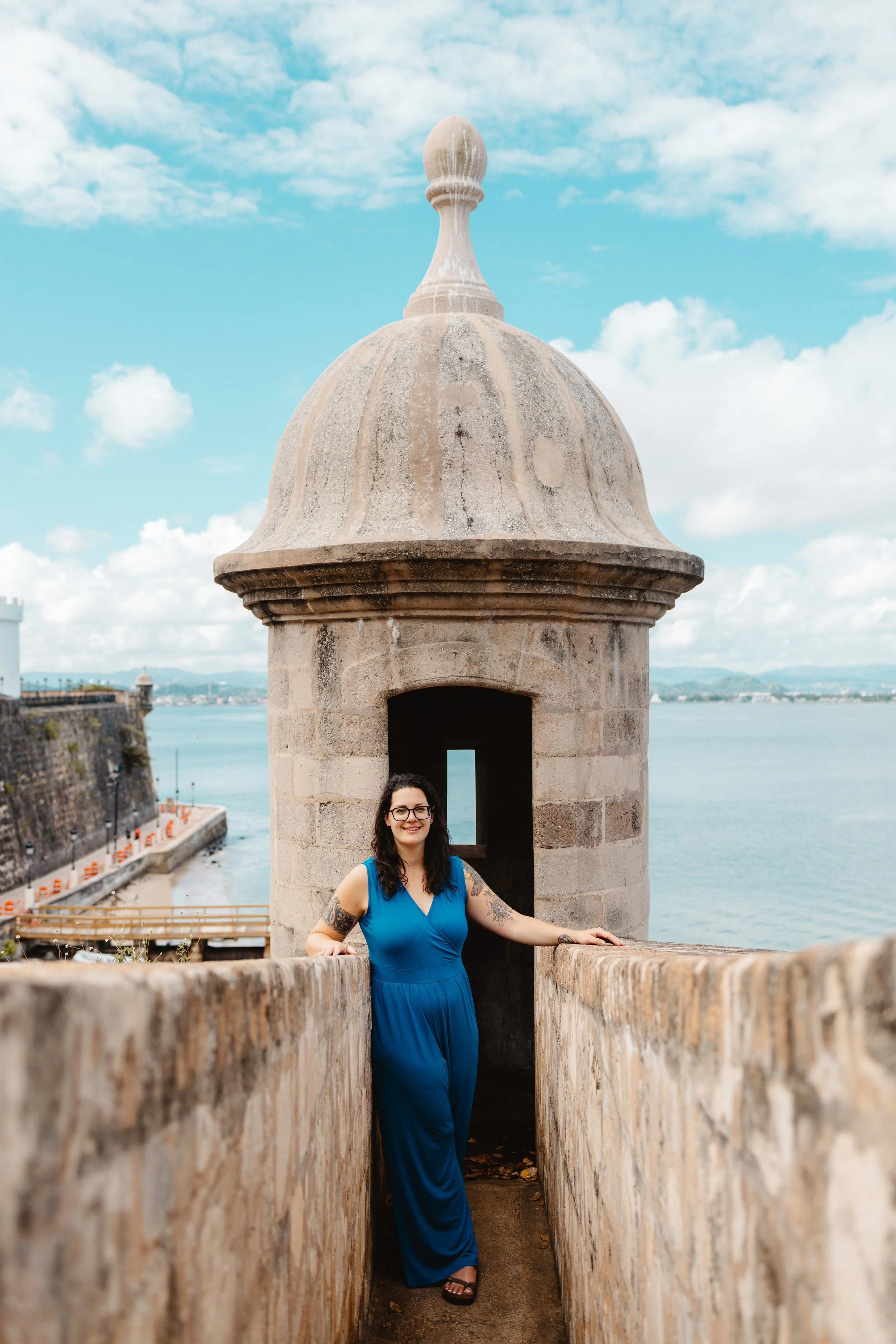 Hannah Clemente of Clemente Travels Virtuoso Advisor in a blue dress standing inside a historic stone lookout tower overlooking water and sky.