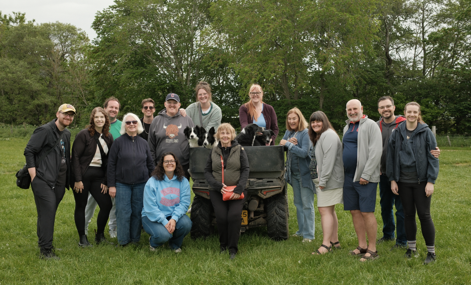 A group of diverse people gathered outdoors on a grassy field with trees in the background, some with dogs and a small ATV.