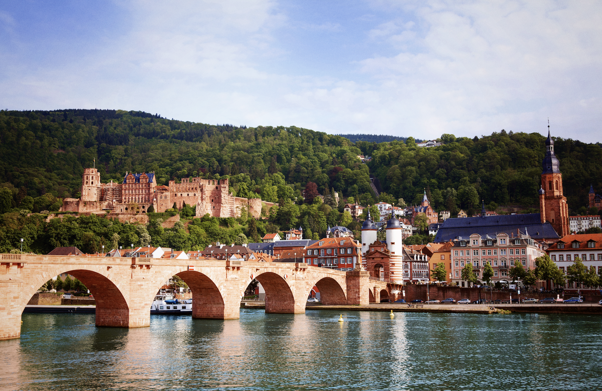 A historic city with a bridge over a river, surrounded by green hills, featuring a castle, a church with a tall spire, and traditional buildings.