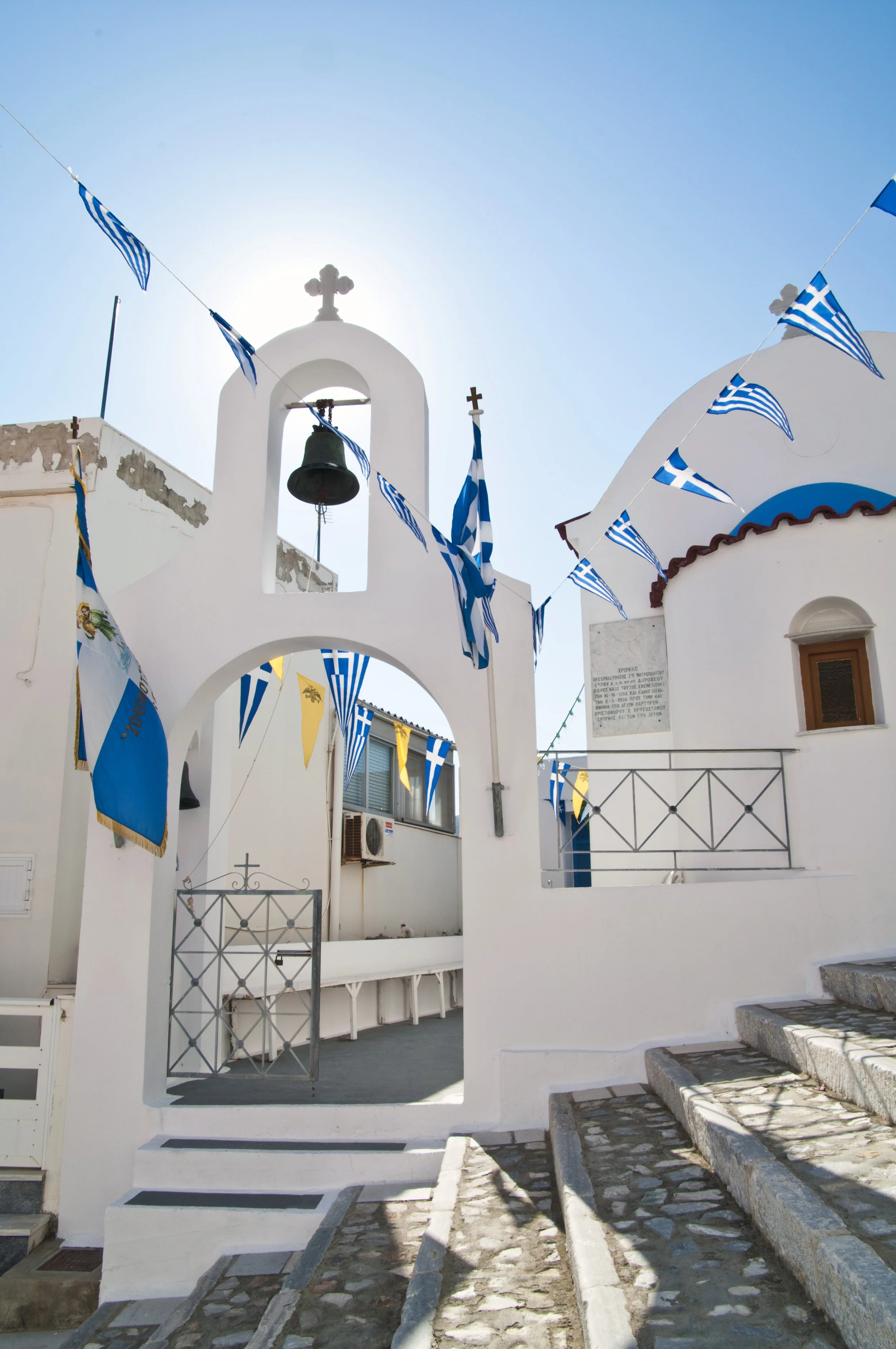 White church with a bell tower, decorated with Greek flags and banners, on a sunny day with a clear blue sky.