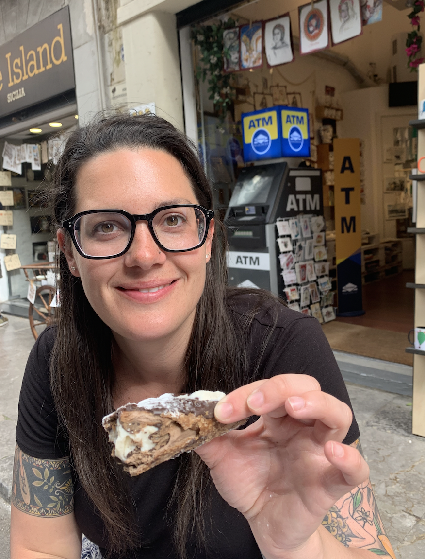 Hannah Clemente a Virtuoso Travel Advisor with long dark hair, glasses, and a black shirt smiling and holding a slice of chocolate pie with whipped cream outside a shop in Sicily Italy. Italy travel specialist. Massillon, Ohio travel advisor