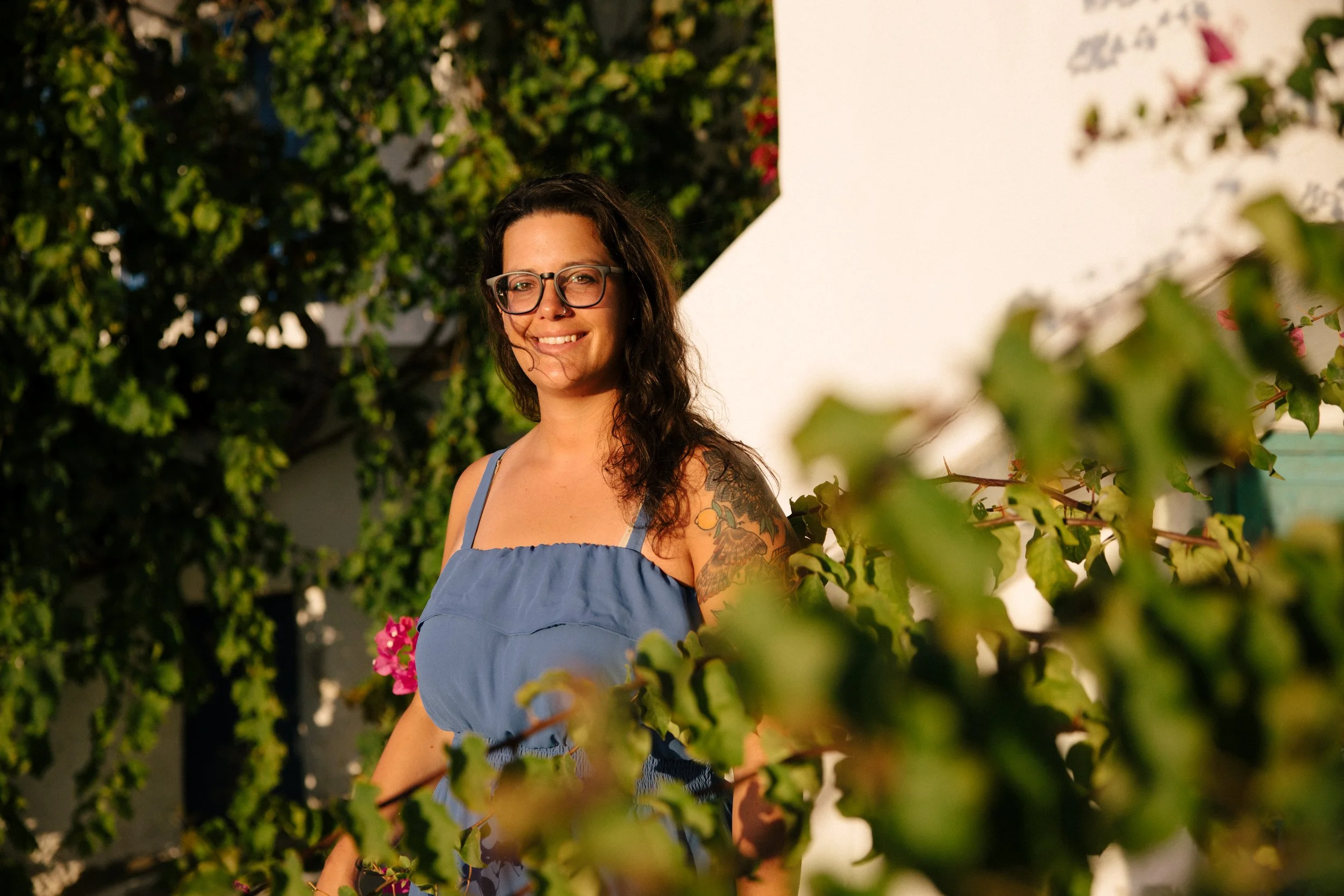 Hannah Clemente of Clemente Travels a Virtuoso Travel Advisor with long dark hair and glasses smiling outdoors, wearing a blue sleeveless top with a tattoo on her left arm, surrounded by green plants and pink flowers.