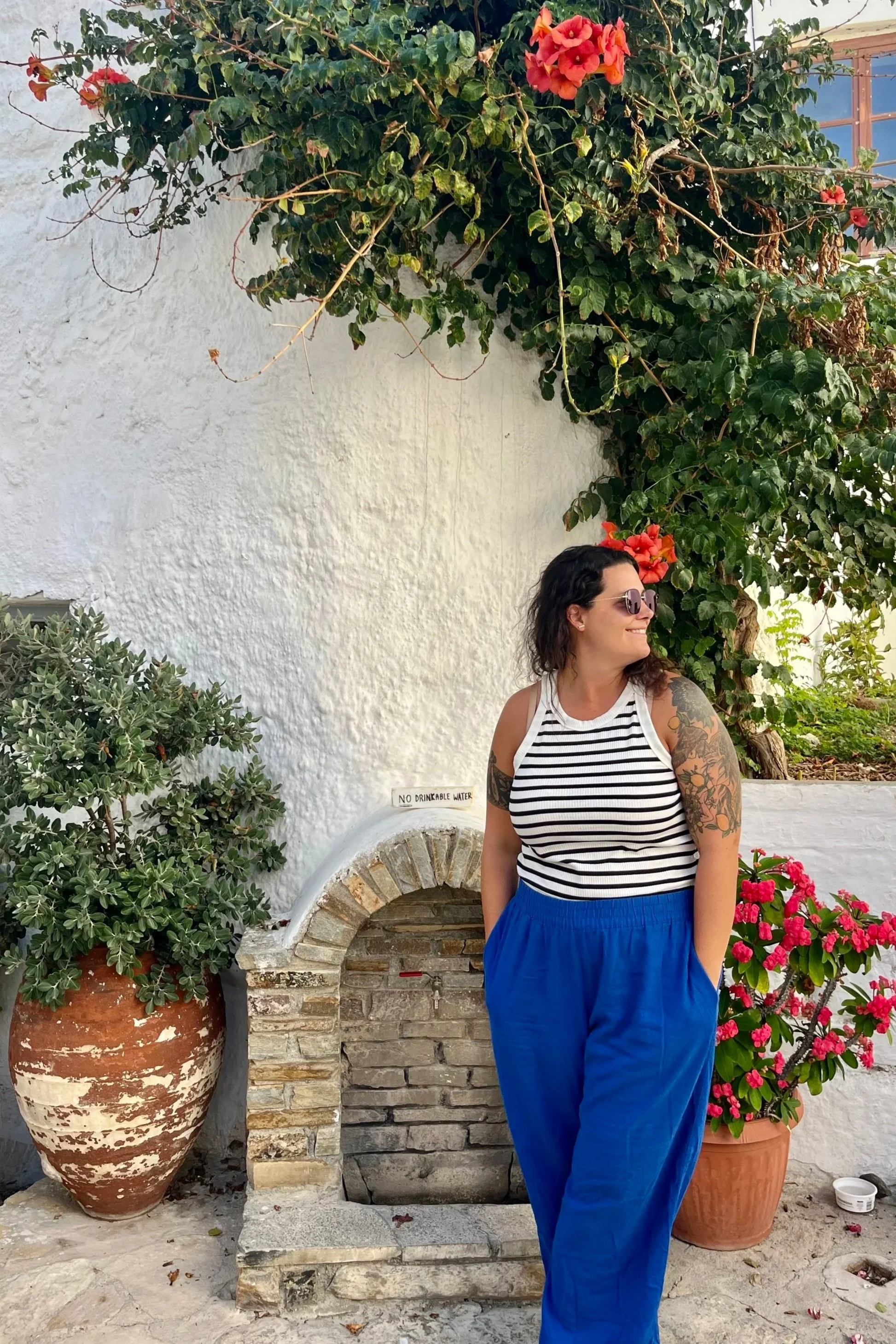 A woman with tattoos on her arms, wearing sunglasses, a white and black striped sleeveless top, and blue pants, standing outdoors beside potted plants and a decorative stone fountain with a sign that reads 'No Drinkable Water'. She is smiling and looking to her right, with a white stucco wall and green leafy climbing plants with pink flowers in the background.