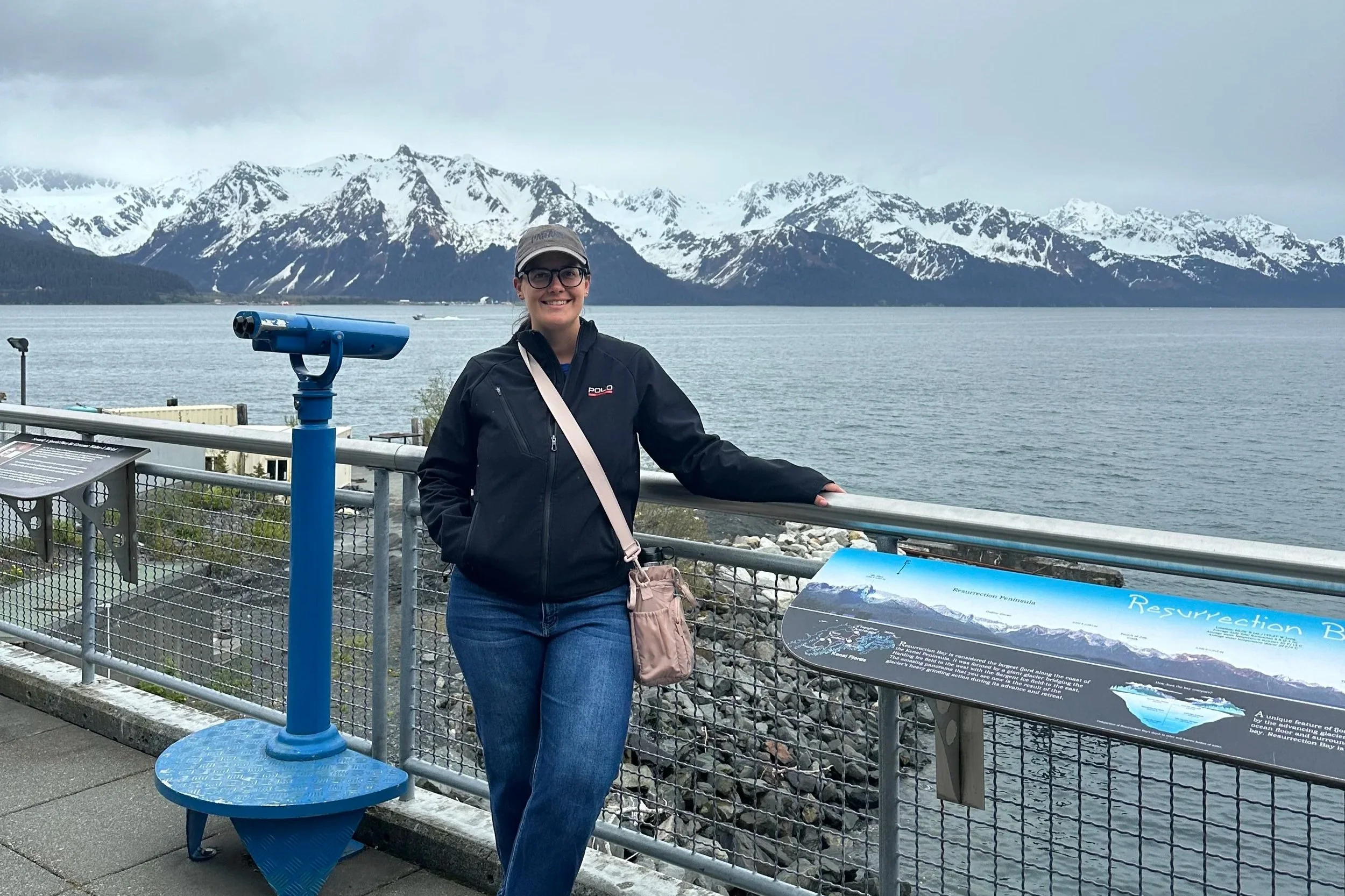 A woman in glasses and a gray cap smiling, standing by a railing with a scenic lake and snow-capped mountains in the background. She is wearing a black jacket and blue jeans, and has a pink bag over her shoulder. There is a blue coin-operated telescope and an informational sign nearby.