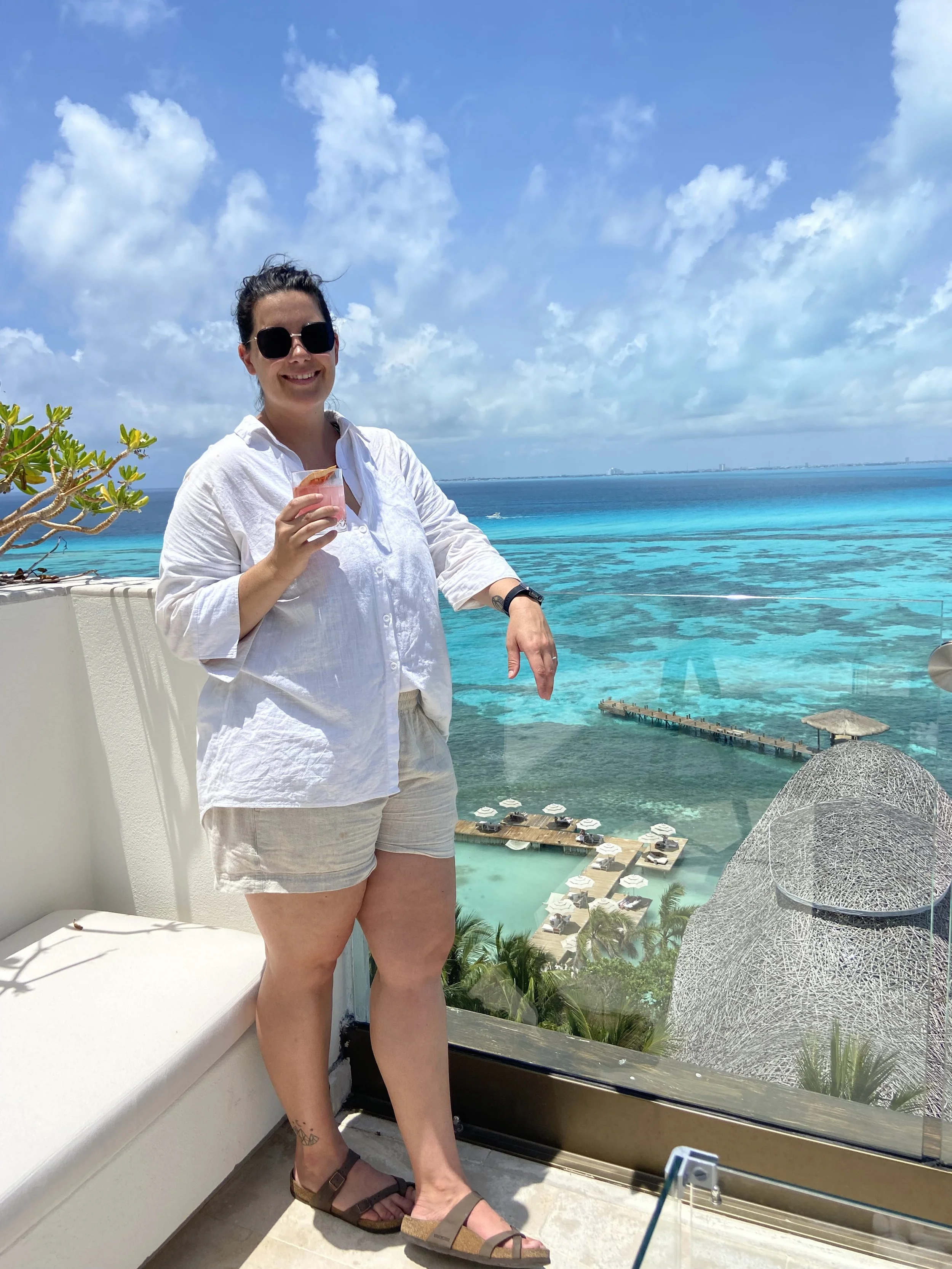 Hannah Clemente of Clemente Travels a Virtuoso Travel Advisor wearing a white shirt and shorts holding a drink, standing on a balcony overlooking a tropical beach with clear blue water, umbrellas, and overwater bavilions under a partly cloudy sky.