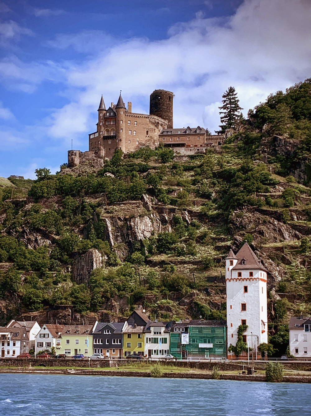 A historic castle atop a green, rocky hillside overlooking colorful houses and a river.