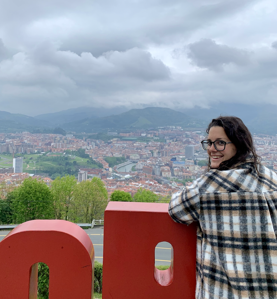 Hannah Clemente of Clemente Travels a Virtuoso Travel Advisor with dark curly hair and glasses smiling while leaning on a red railing overlooking a cityscape with mountains and cloudy sky in the background. Massillon, Ohio travel advisor.