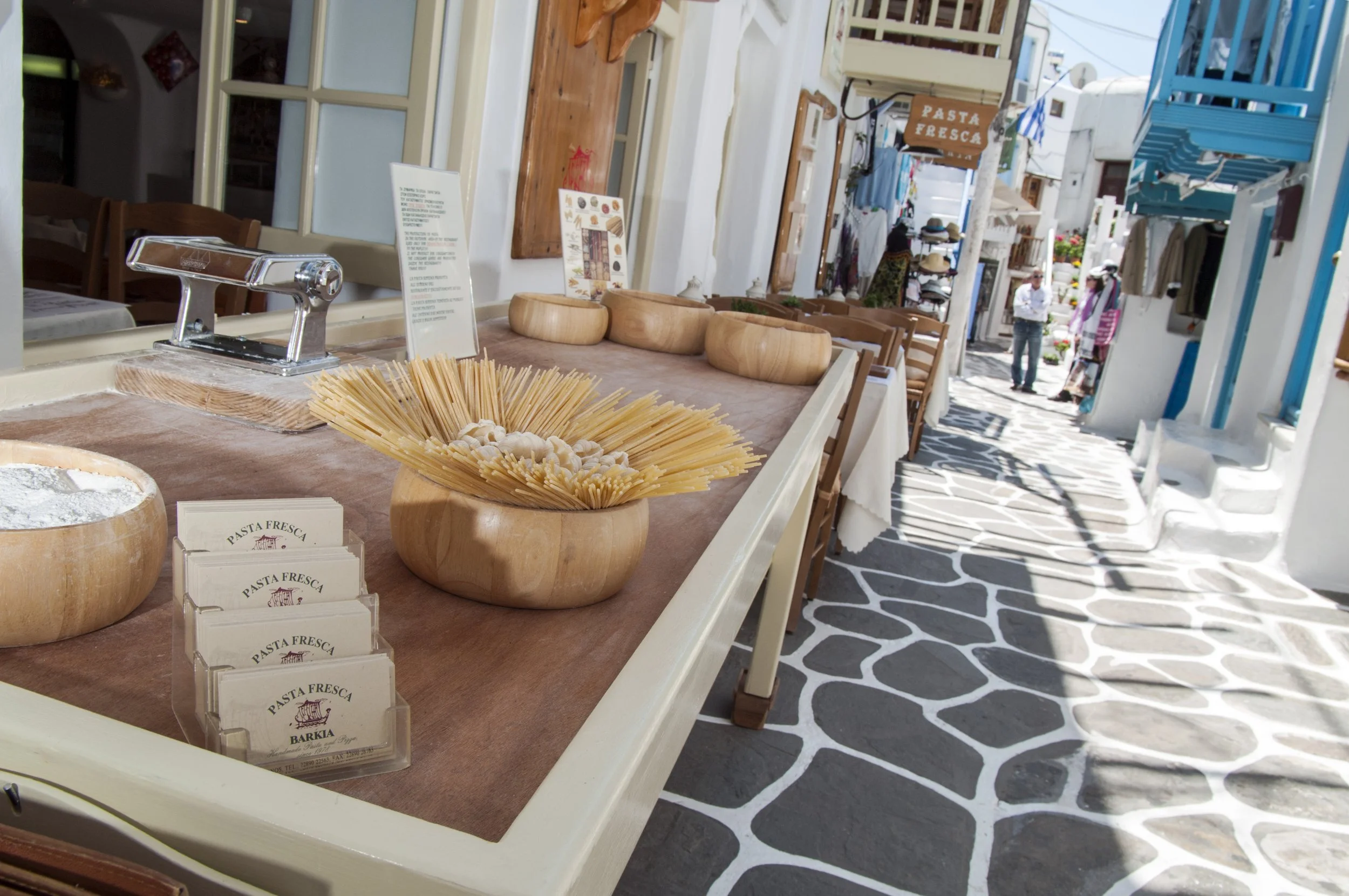 Outdoor cafe with wooden bowls of pasta and a menu on a counter, narrow street with white buildings and blue awnings, sign reading 'Pasta Fresca'.