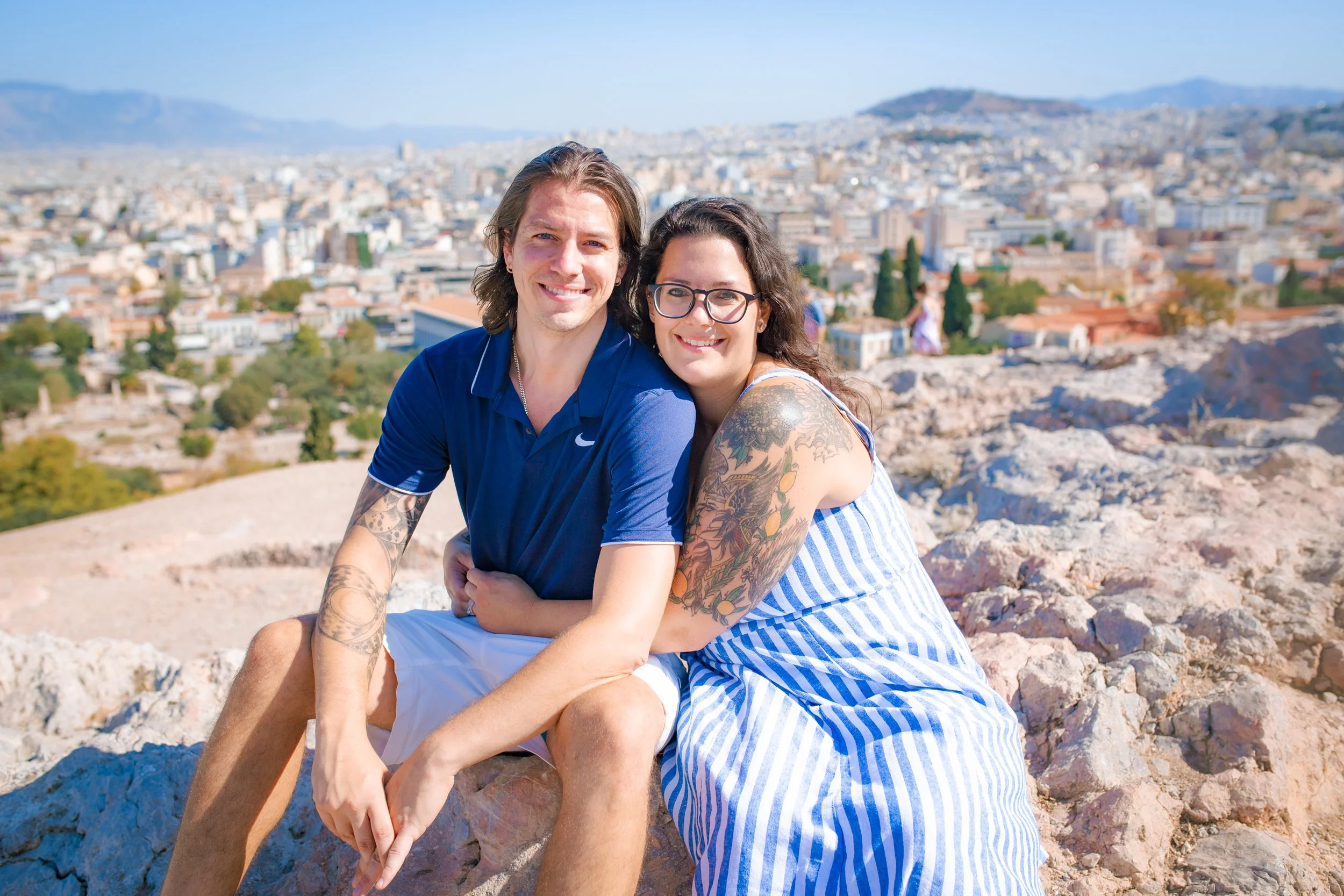 A smiling couple sitting on rocks with a cityscape and mountains in the background on a sunny day.