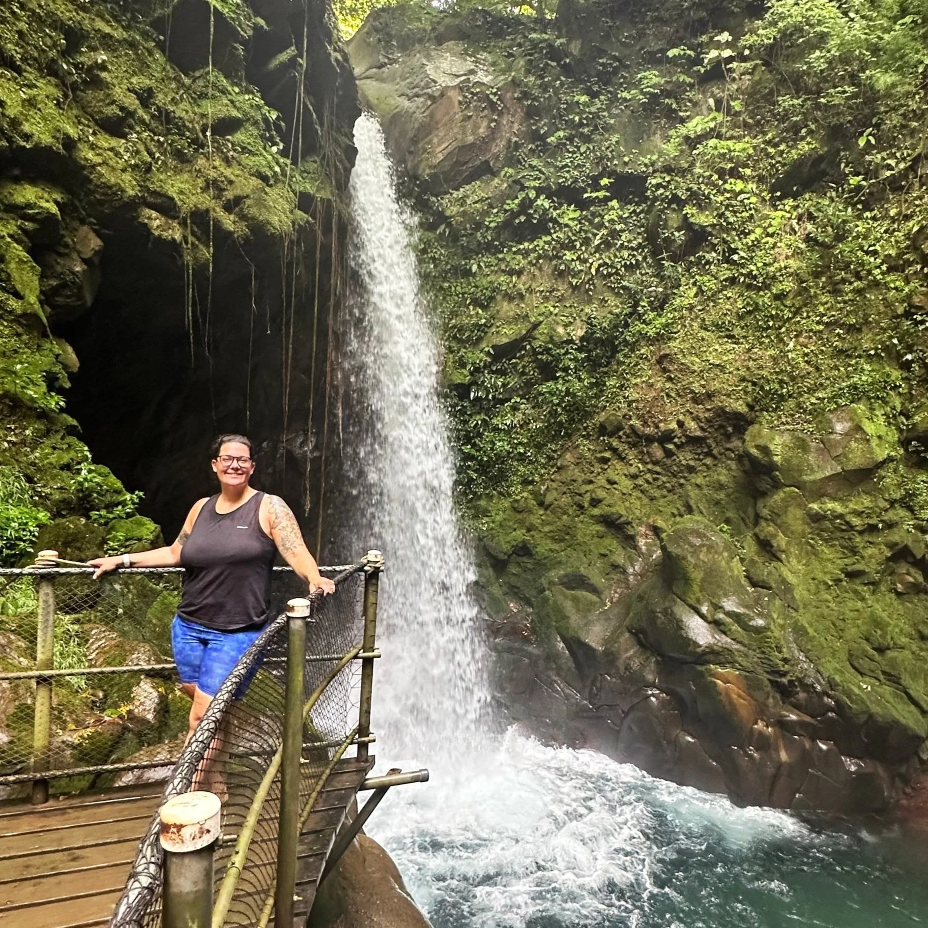 Hannah on a wooden platform in front of a costa rican waterfall wearing black shirt and blue shorts