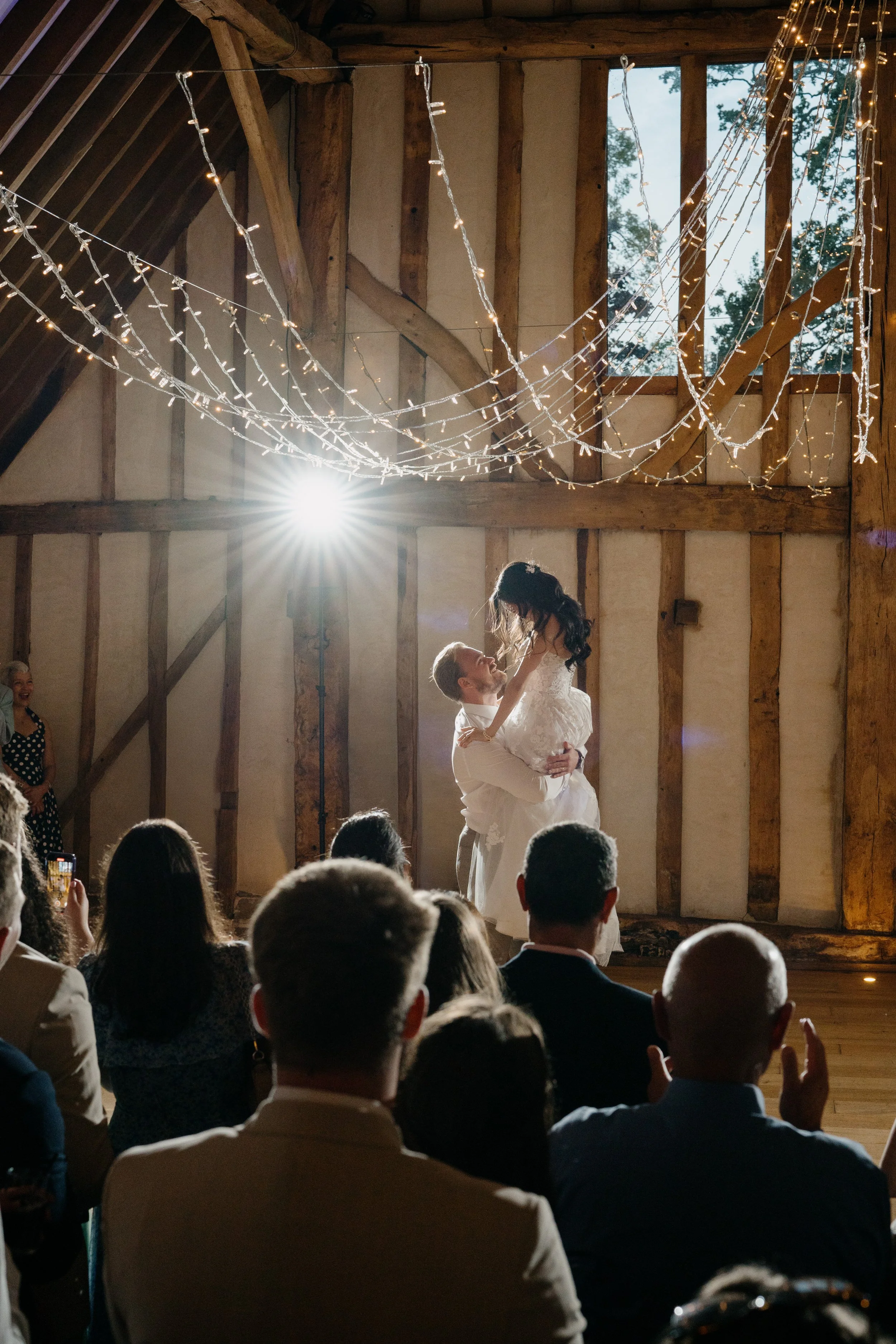 A bride and groom dancing at their wedding reception inside a rustic barn with string lights and a large window, surrounded by seated guests.