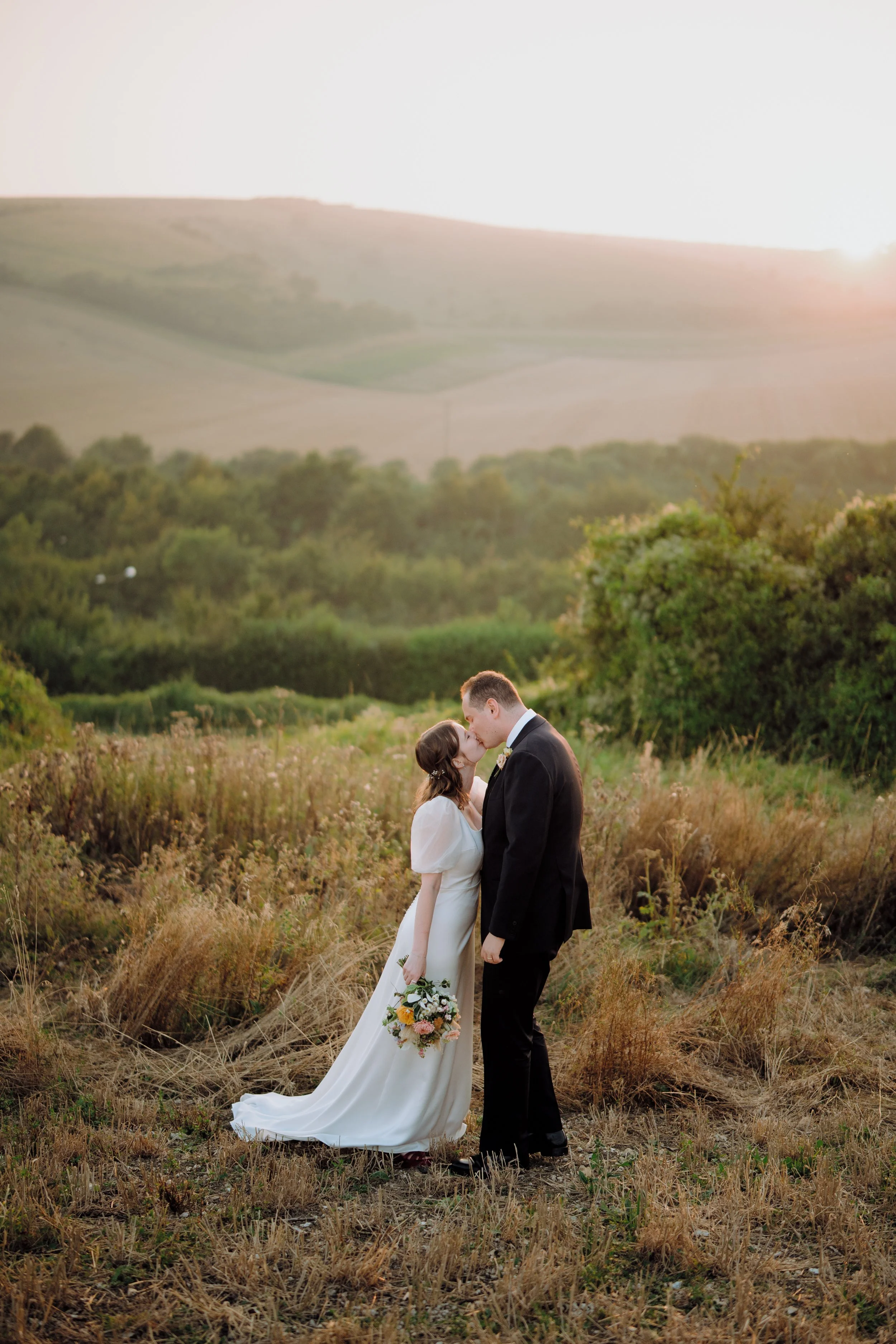 A bride and groom sharing a kiss outdoors during sunset, with a scenic landscape of hills and greenery in the background.