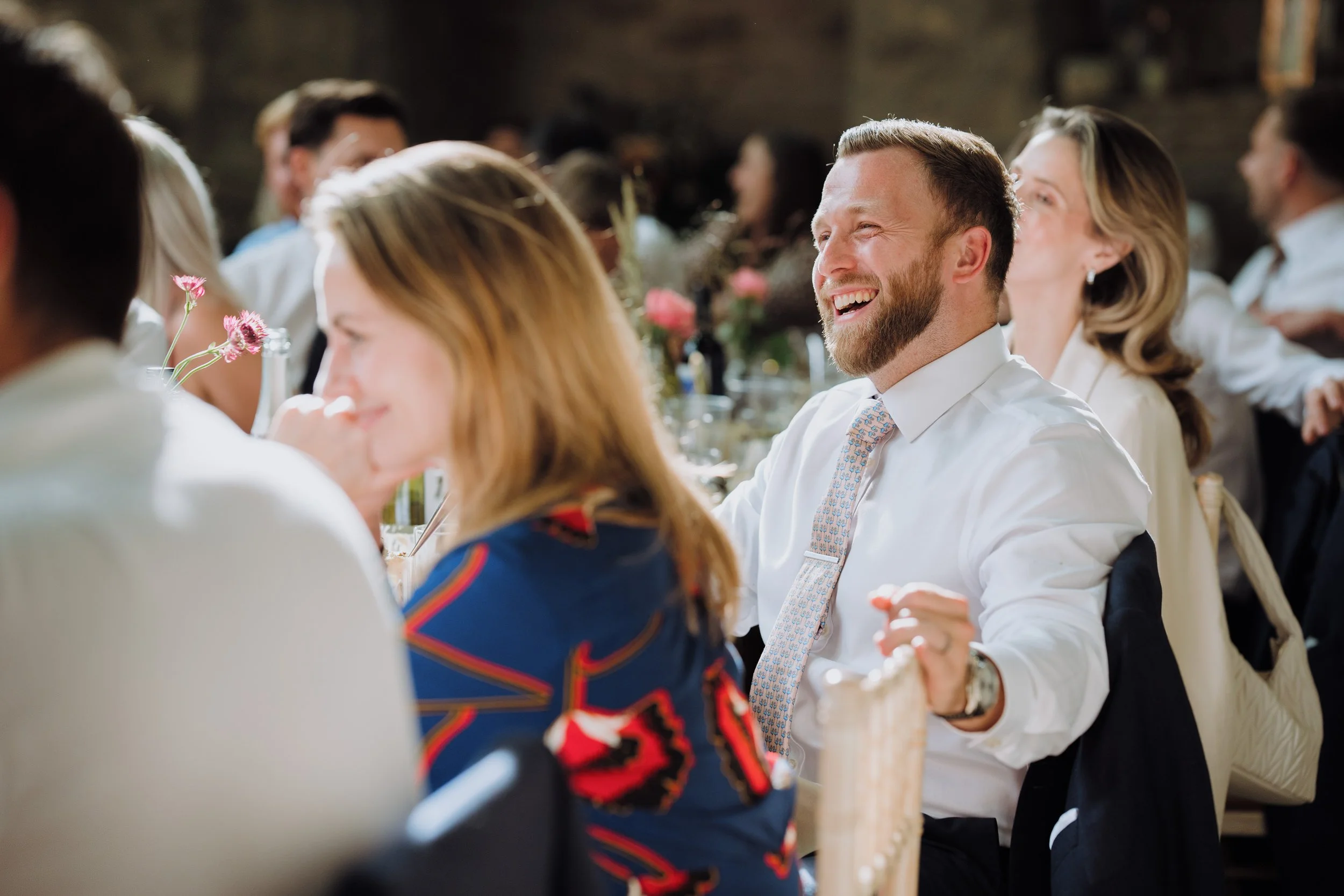 People gathered at a formal event, with a man in a white shirt and tie smiling and laughing, surrounded by women and men seated at tables decorated with flowers and candles.