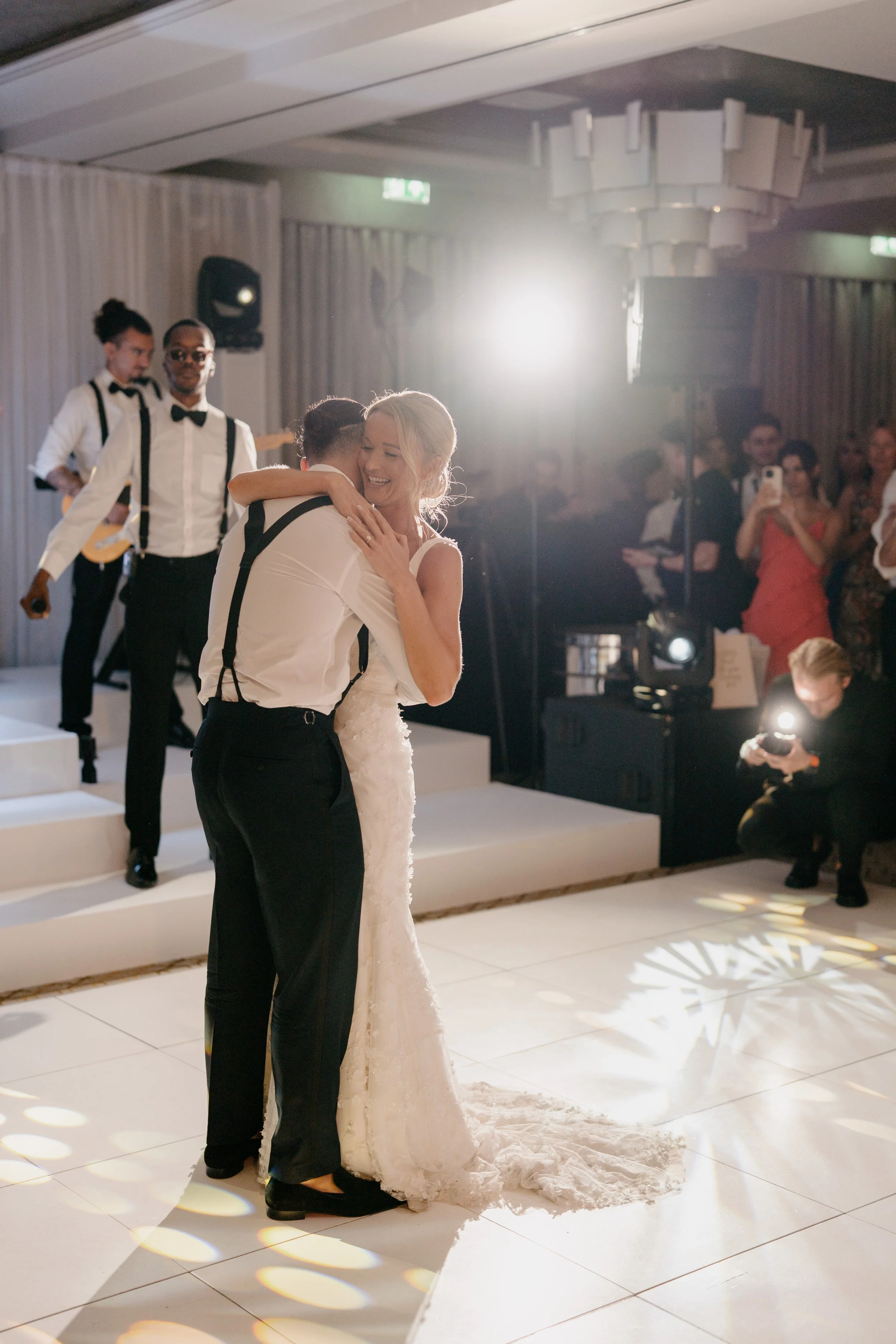 A bride and groom embracing and dancing at their wedding reception. The bride is wearing a white wedding dress, and the groom is dressed in black pants, a white shirt, and suspenders. There are wedding guests and musicians in the background, with pro