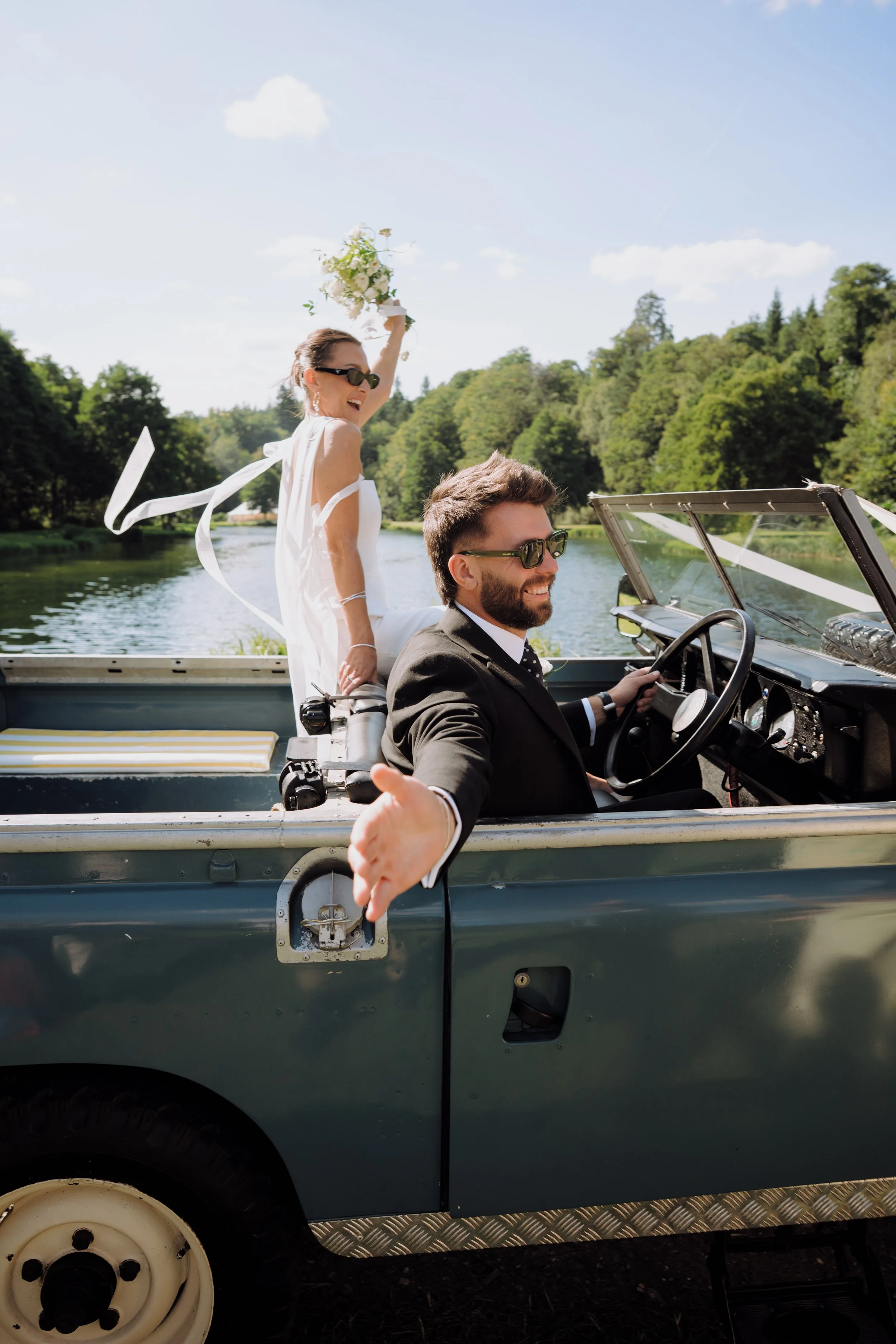 A groom in a tuxedo driving a Land Rover with a woman in a white dress sitting on the edge of the vehicle, holding a bouquet of flowers, on a river surrounded by green trees on a sunny day.