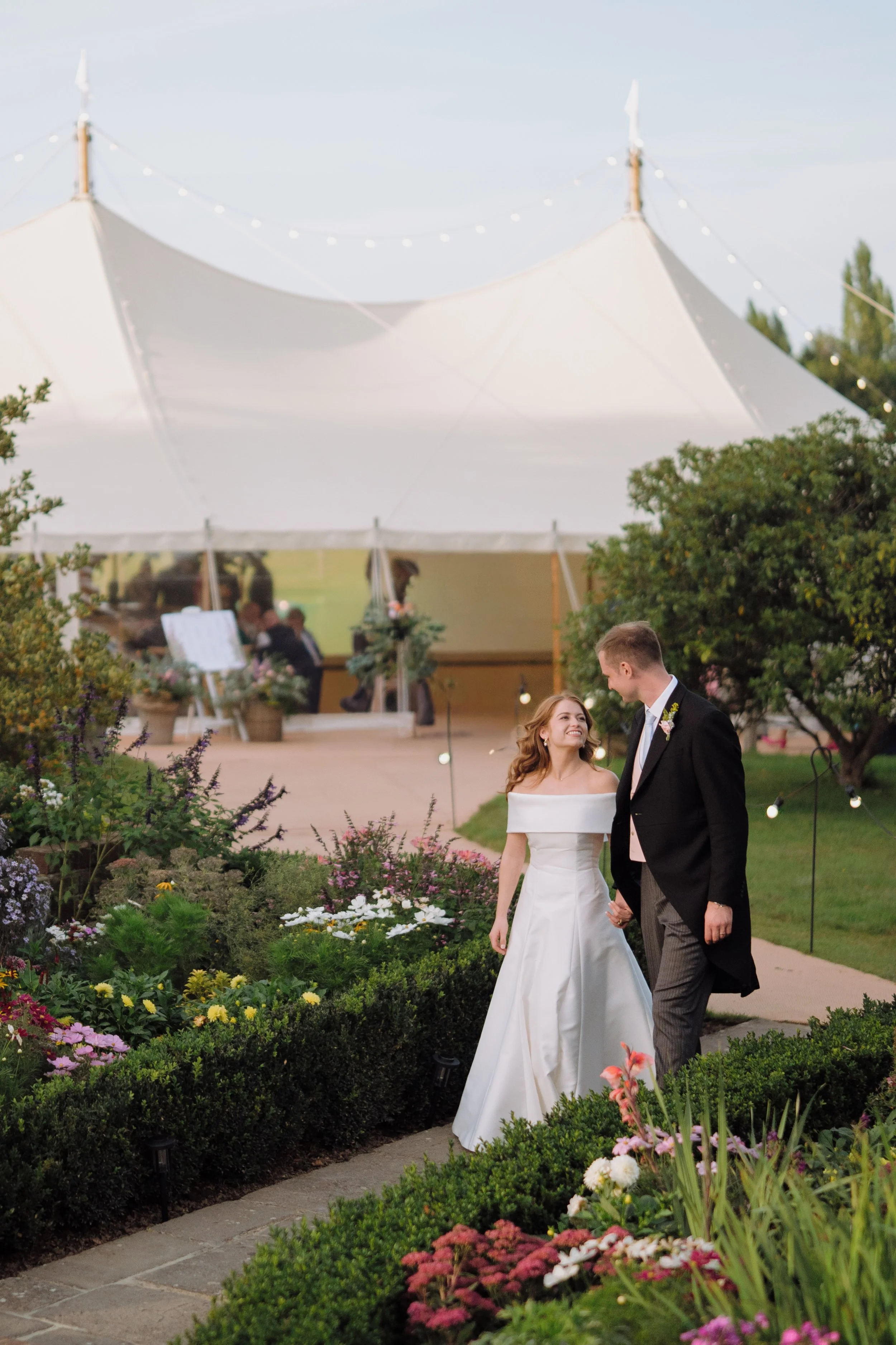 A bride and groom holding hands and looking at each other while walking through a Cotswold garden with colorful flowers during their wedding celebration at an outdoor venue with a marquee in the background.
