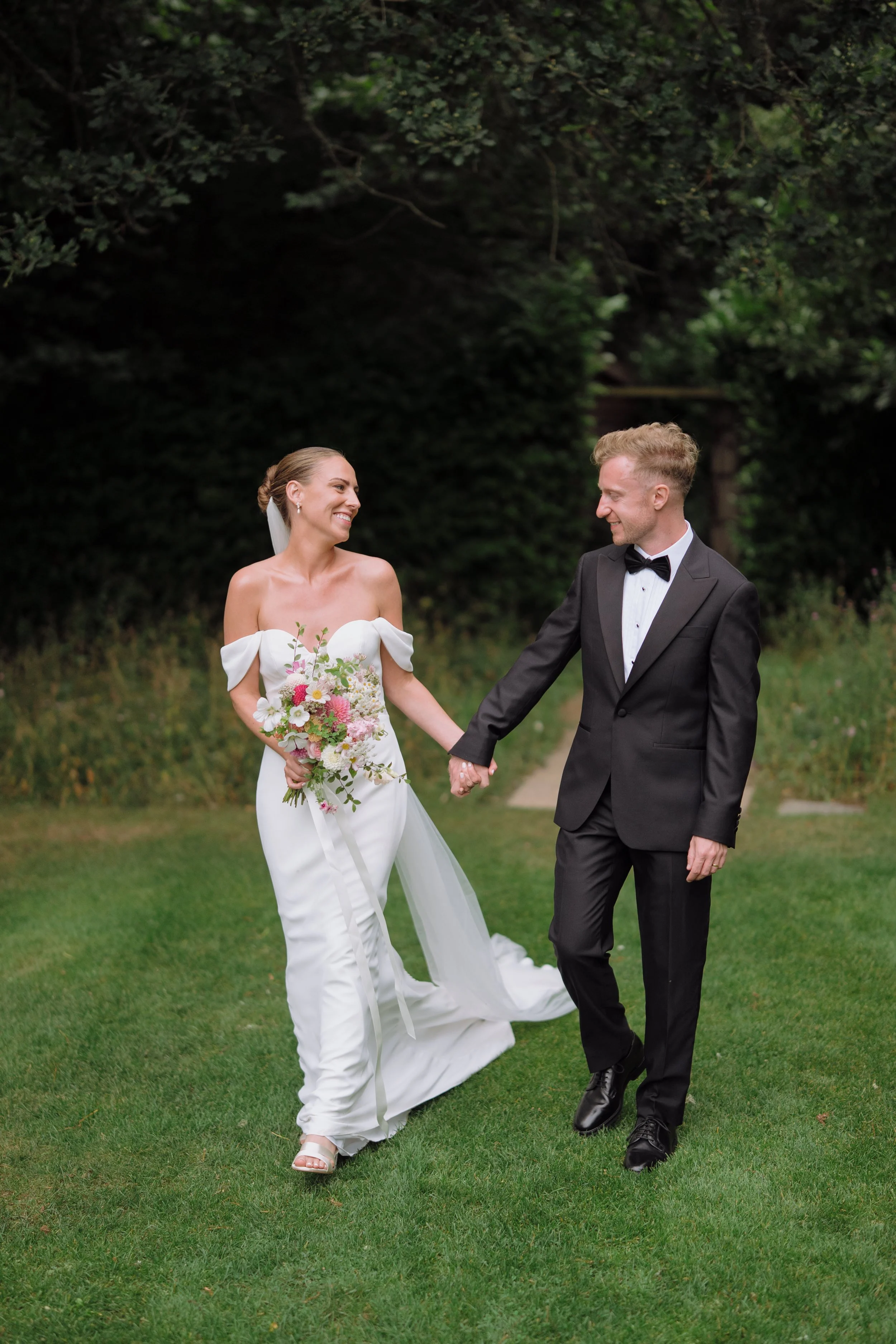 A bride and groom holding hands and smiling at each other outdoors on a grassy area with trees in the background.