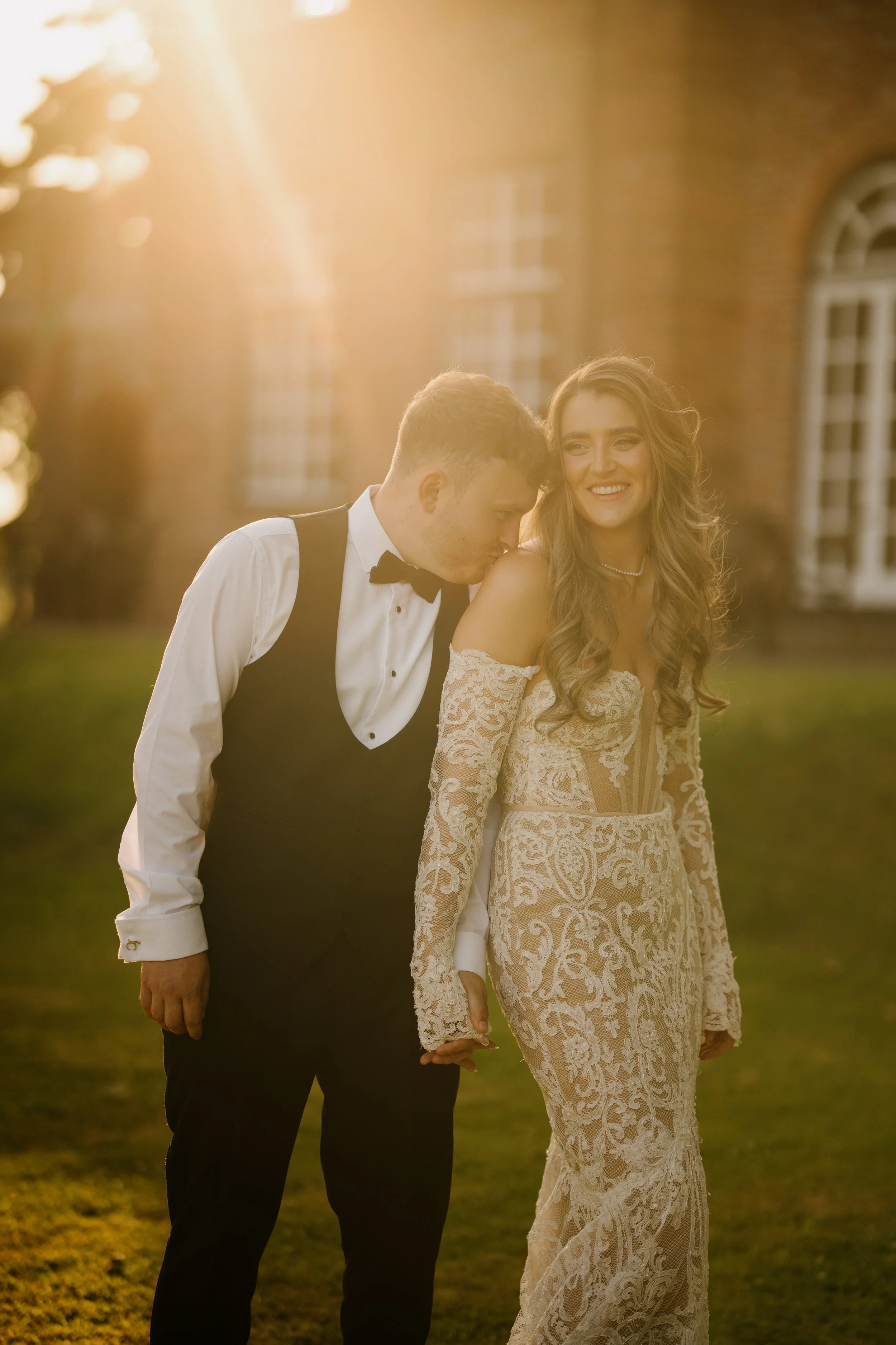 A bride and groom walking outdoors in elegant wedding attire at sunset, smiling and holding hands.