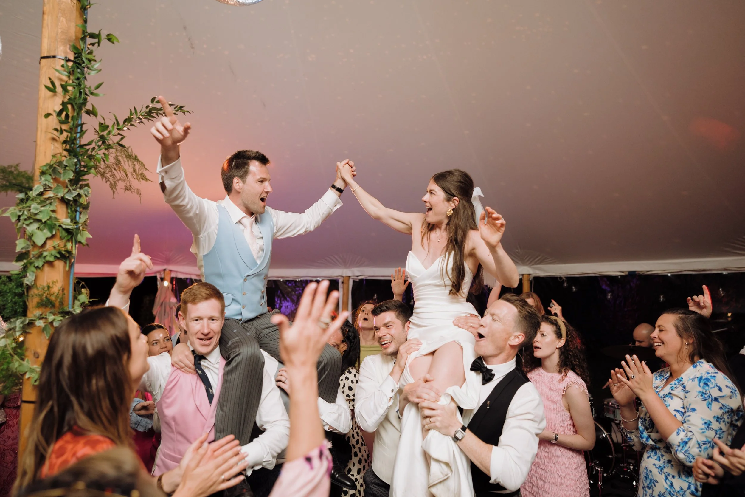 Group of people celebrating at a marquee wedding reception, with the bride and groom riding on the shoulders of friends as others cheer and clap.