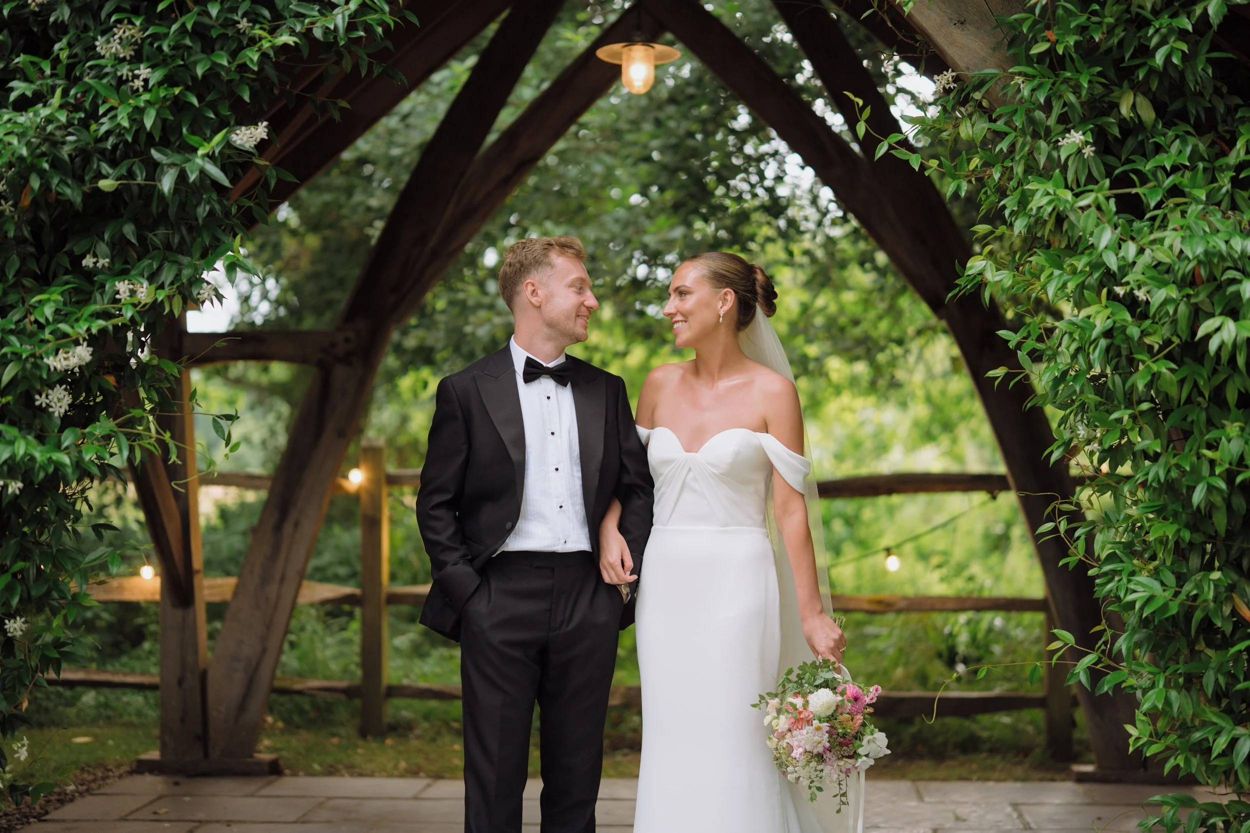 A bride and groom standing under a wooden arch covered with greenery and flowers, smiling at each other, outdoors during the day. The bride wears a white off-shoulder wedding dress holding a bouquet, and the groom wears a black tuxedo with a bow tie.