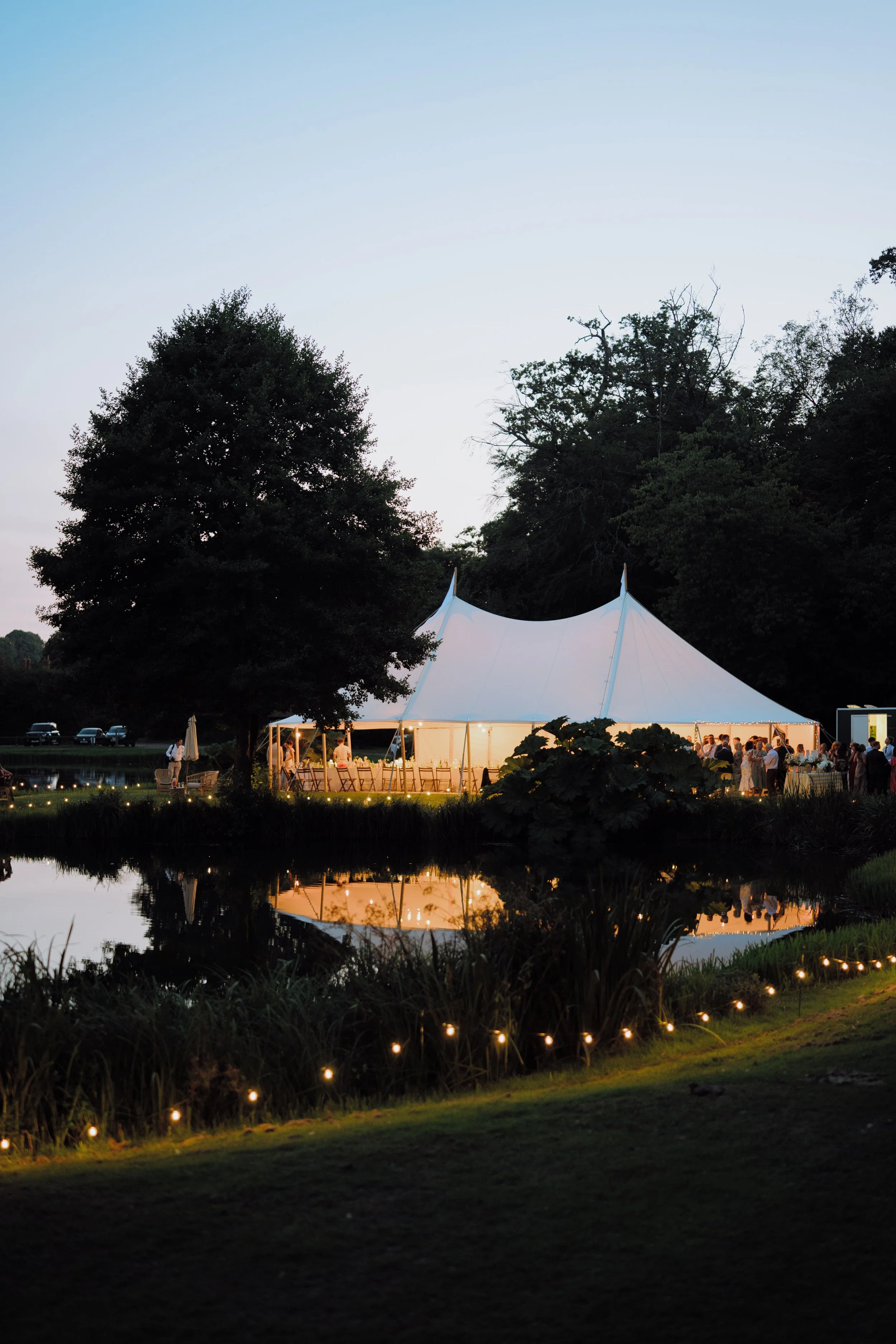 Outdoor wedding reception at twilight with a large white tent illuminated from inside, surrounded by trees, a pond reflecting the tent, and string lights along the grassy area.