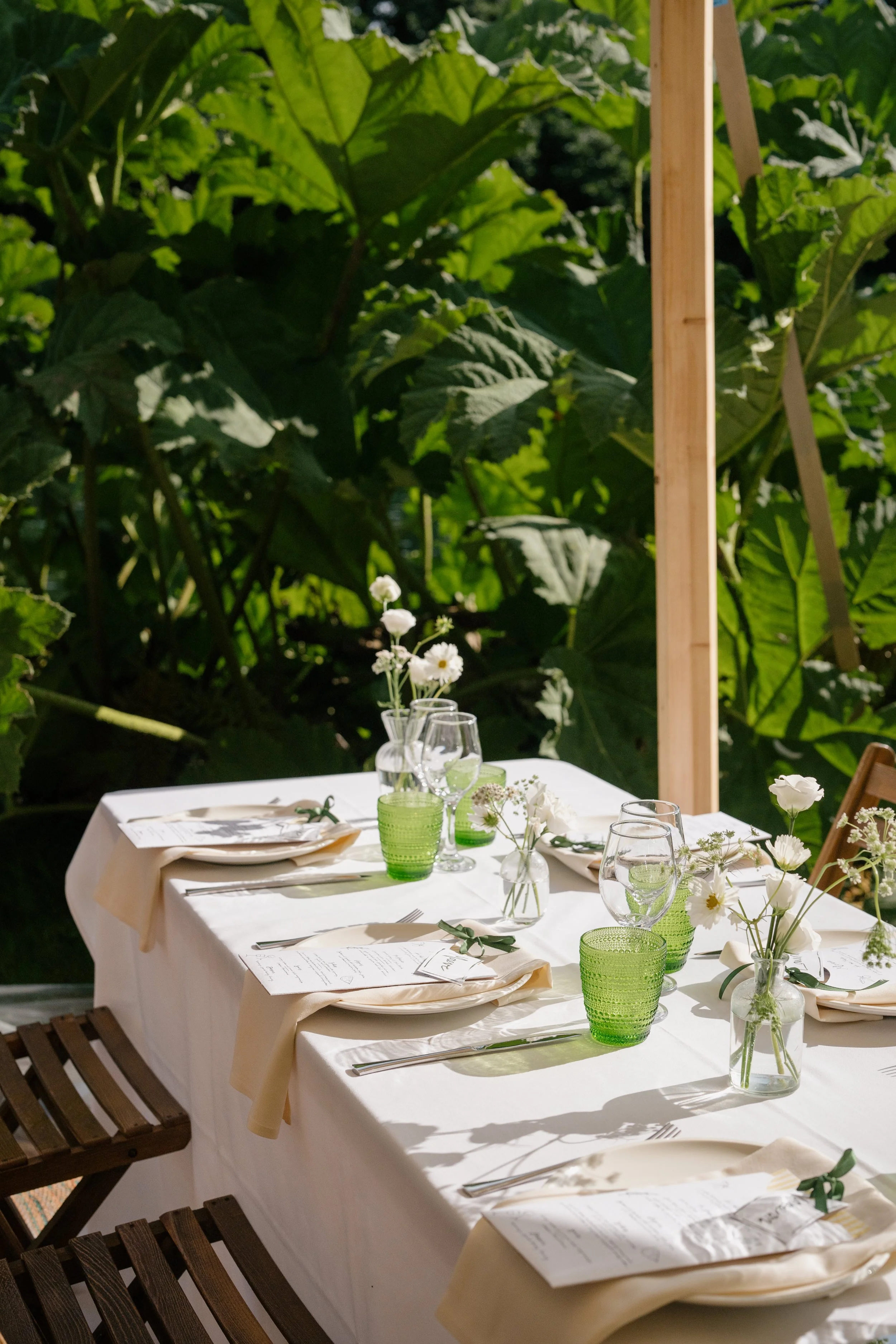 Outdoor dining table set with white tablecloth, green glasses, clear wine glasses, and small white floral centerpieces on a sunny day surrounded by large green leaves.