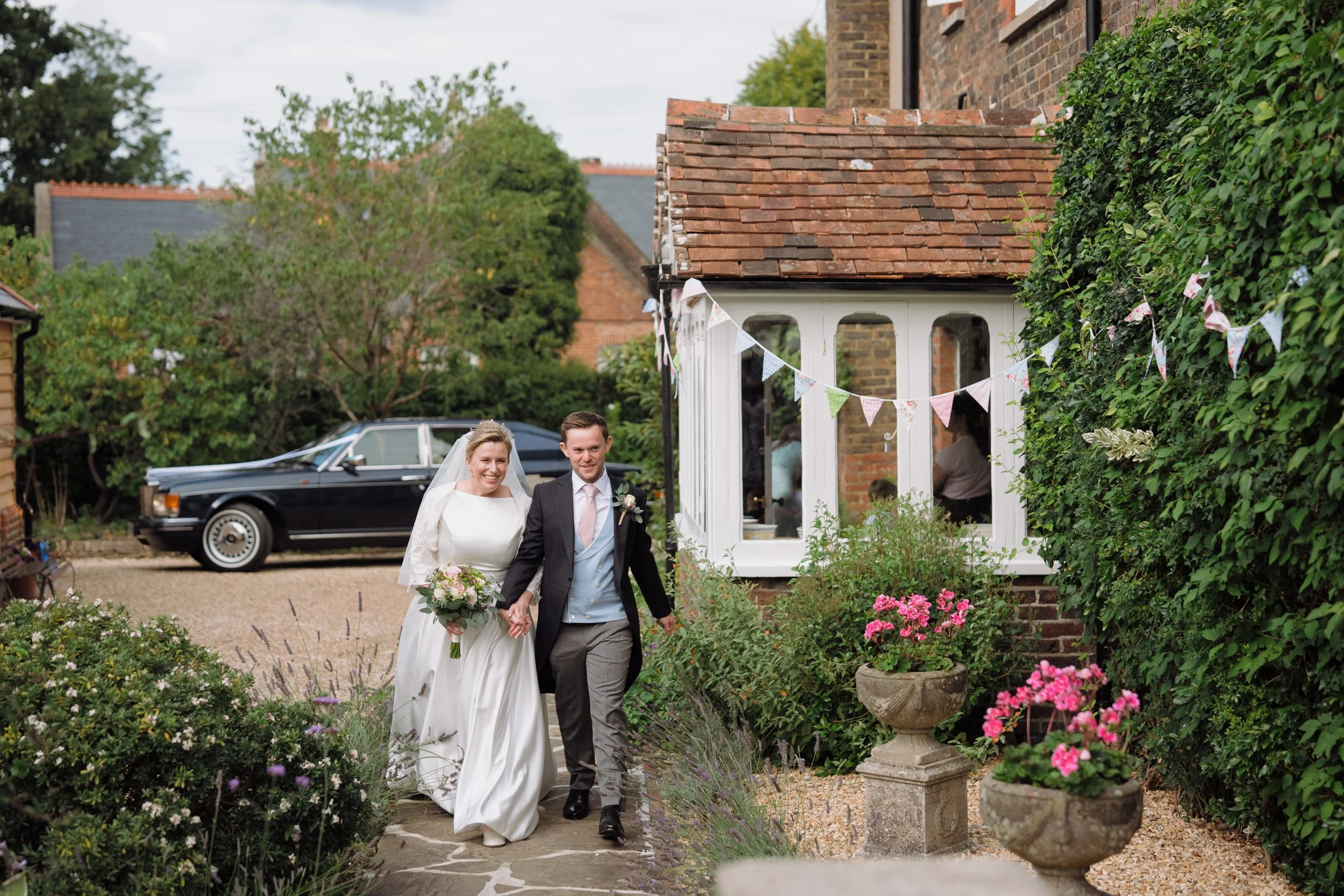 Bride and groom walking hand in hand outside a house decorated with bunting, surrounded by flowers and greenery.