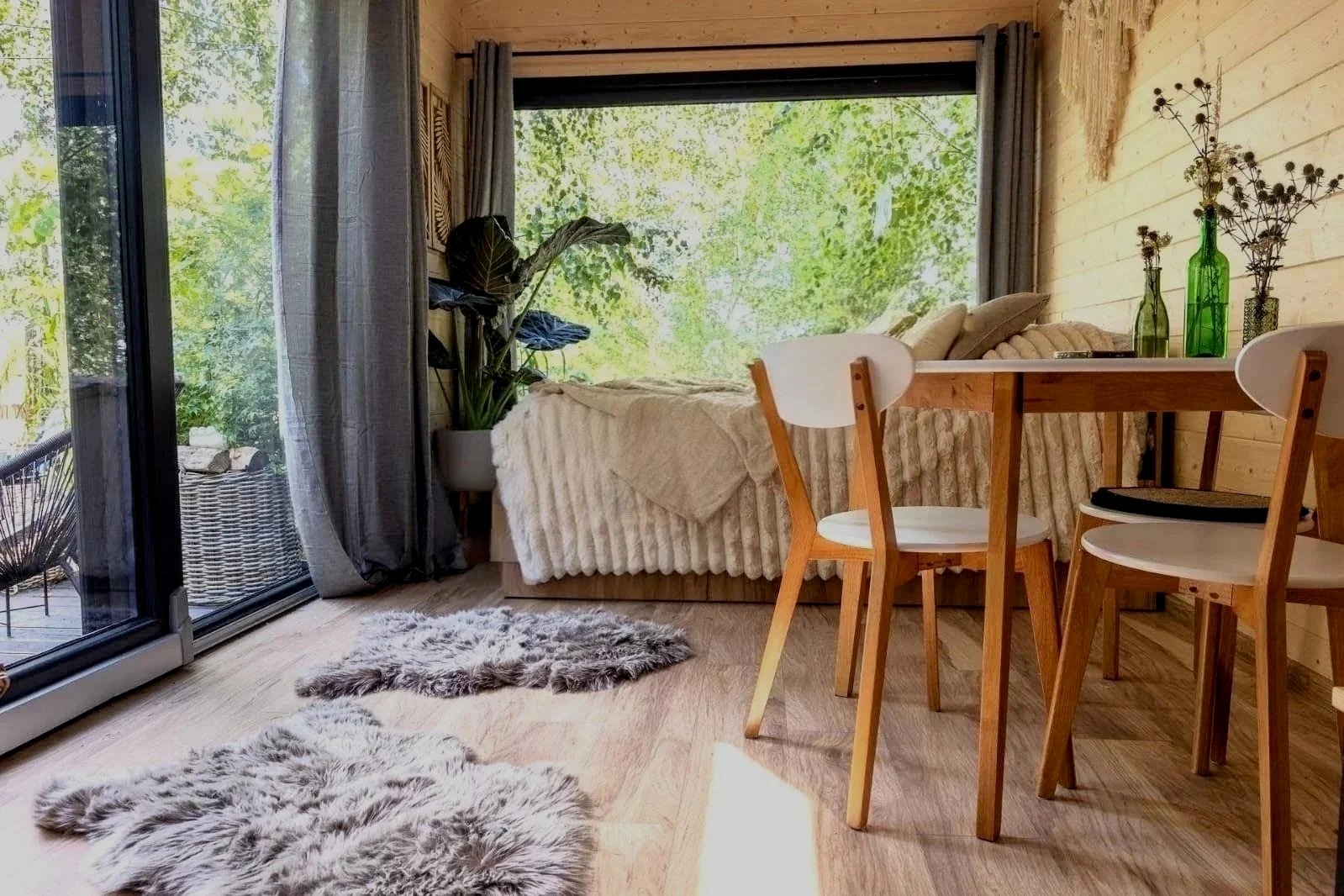 Wohnzimmer mit großen Fenstern, Holzwänden, einem Bett neben der Fenster und einem Essbereich mit Holzstühlen und einer weißen Tischplatte, mehrere grüne Vasen mit Blumen auf dem Tisch, Fellteppiche auf dem Holzboden.