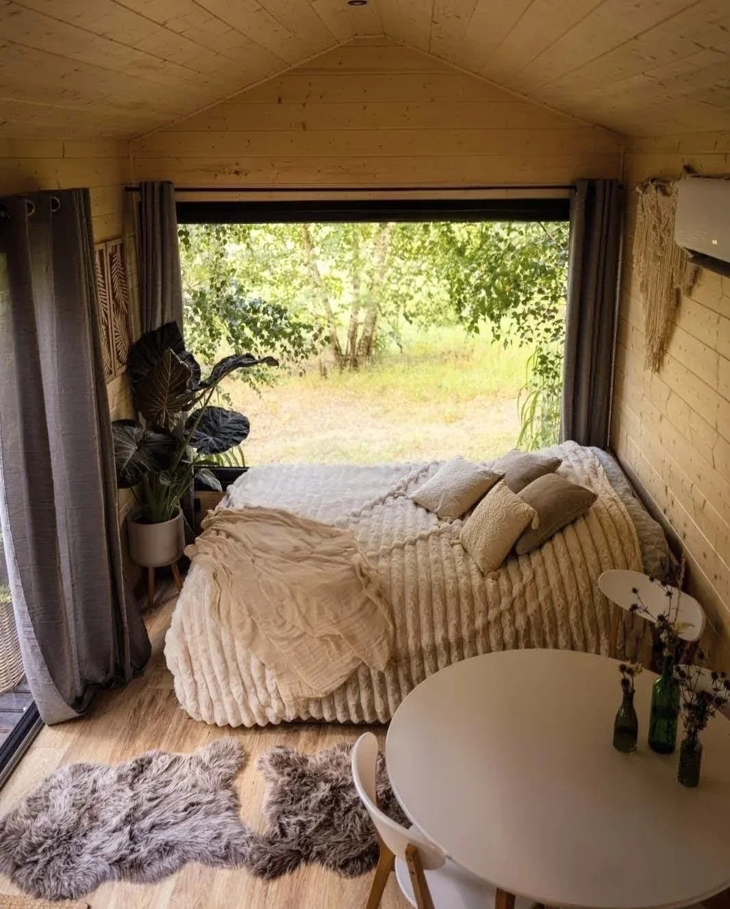 Ein gemütliches Schlafzimmer mit einem Bett, das mit kuscheligen Decken und Kissen bedeckt ist, vor einem großen Fenster mit Blick ins Grüne. Es gibt Pflanzen, ein runder Tisch mit Vasen und Stühlen, sowie einen Teppich aus Fell.