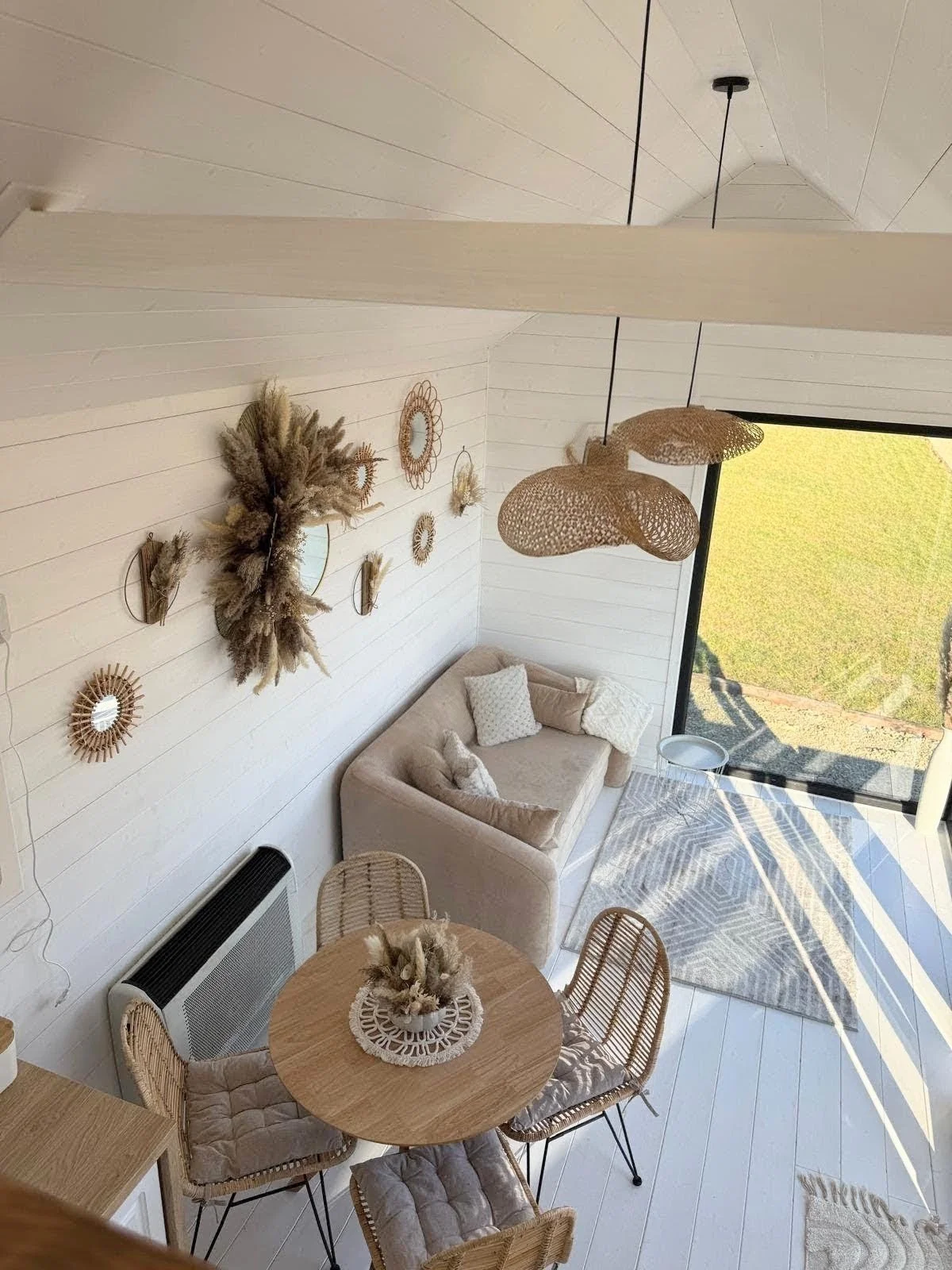 Heller, moderner Wohnraum mit weißen Wänden, Kissen auf einem Sofa, dekorativen Wandspiegeln und Pflanzen, großer Fensterfront mit Blick auf eine grüne Wiese, runde Holztisch mit Korbstühlen, Pendelleuchten aus Holzdeko.